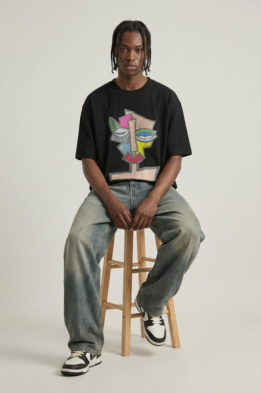 Person wearing a black t-shirt with a colorful graphic design, sitting on a wooden stool against a plain background.