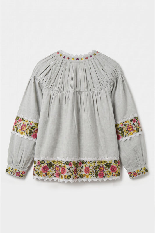 A light gray long-sleeve blouse with a pleated bodice, floral embroidered cuffs, and a matching floral hem, shown against a plain light gray background.
