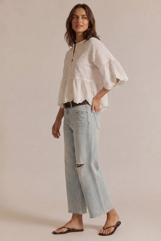 A woman modeling a white long-sleeve blouse with ruffled bell sleeves and a scalloped hem, paired with light-wash cropped jeans and dark brown sandals, against a solid beige studio background.