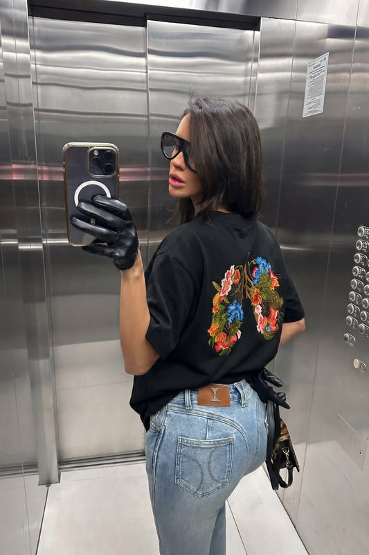 Person taking a mirror selfie in an elevator wearing a black t-shirt with colorful designs and light blue jeans.