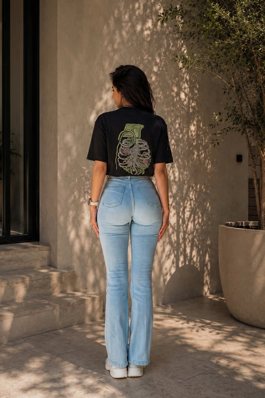 Person wearing a black t-shirt with an embroidery design and light blue jeans standing against a beige wall.