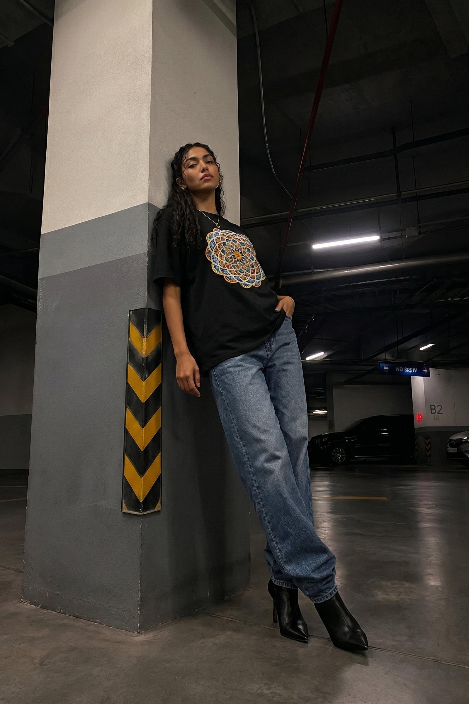 Person wearing a black t-shirt with a colorful embroidery design and blue jeans standing in a parking garage.