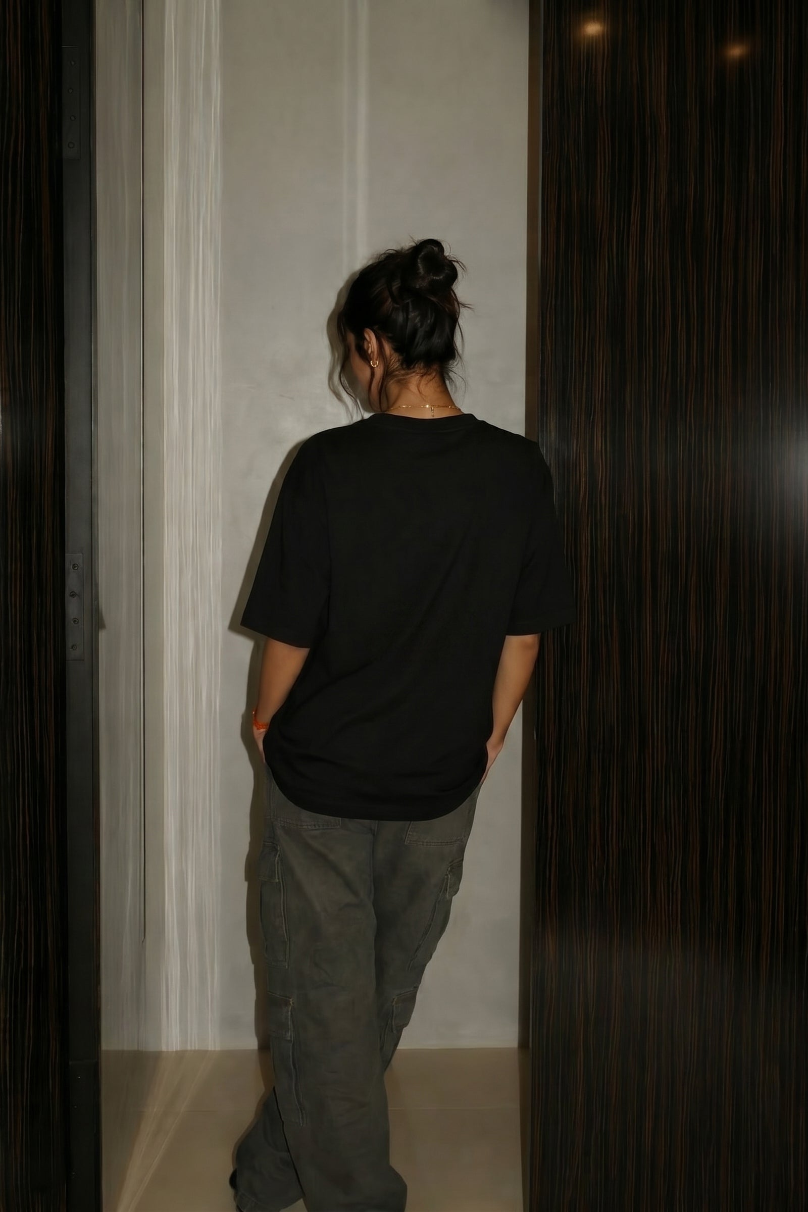 Person wearing a black t-shirt and gray pants standing against a plain wall.