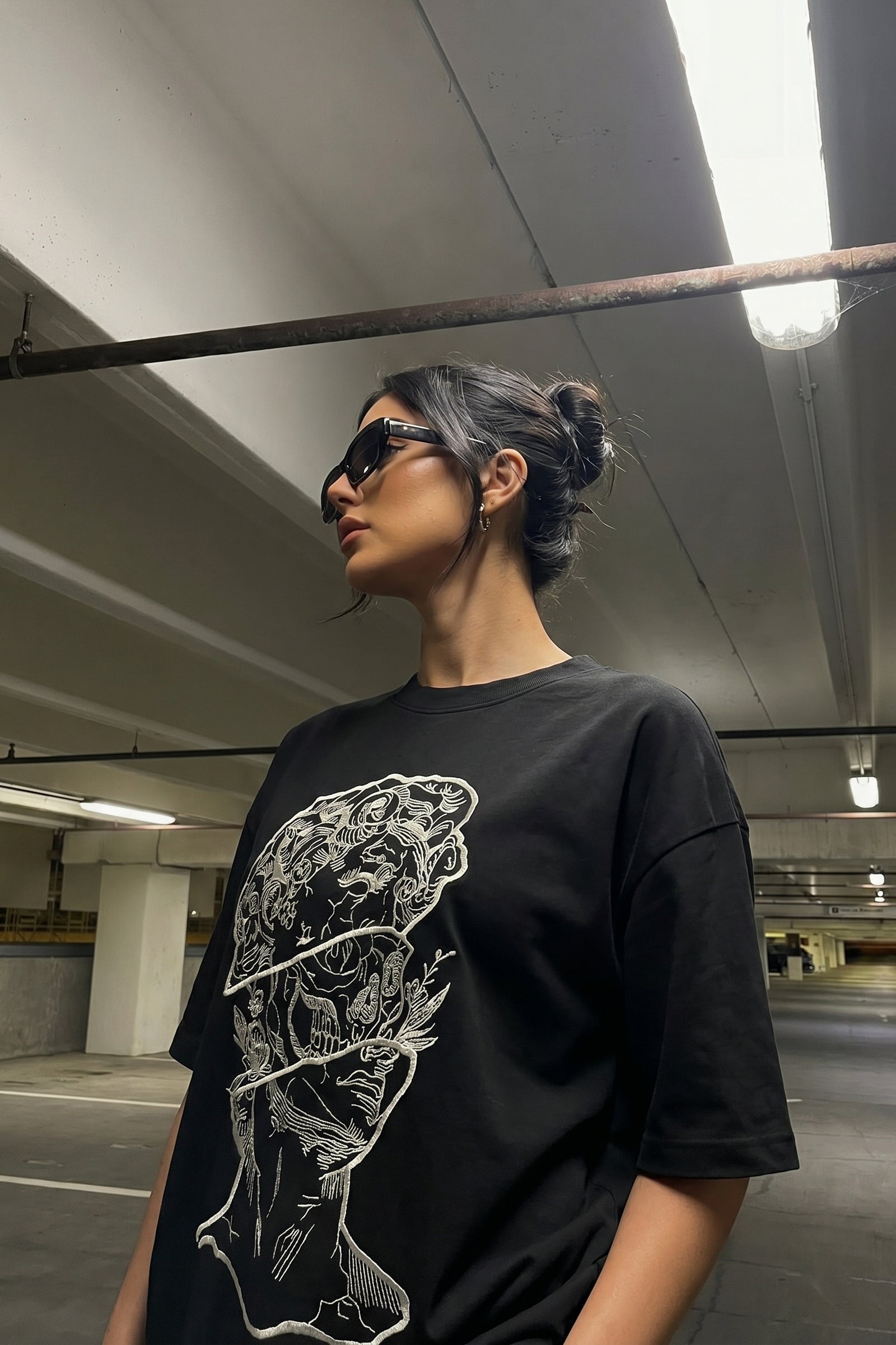 Person wearing a black t-shirt with a embroidery design and gray pants in an underground parking lot.