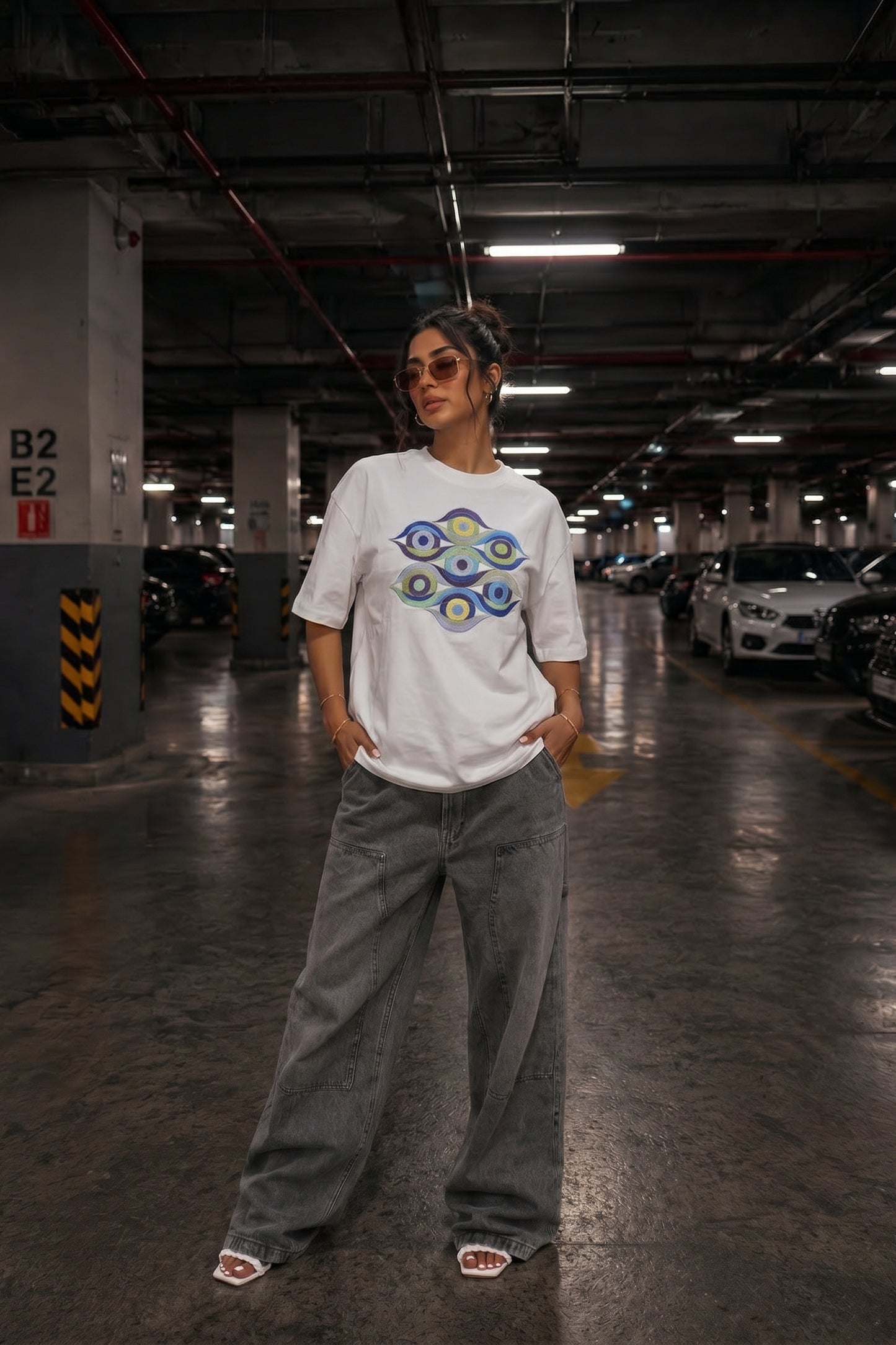 Person wearing a white t-shirt with a colorful embroidery design and gray pants in a parking garage.