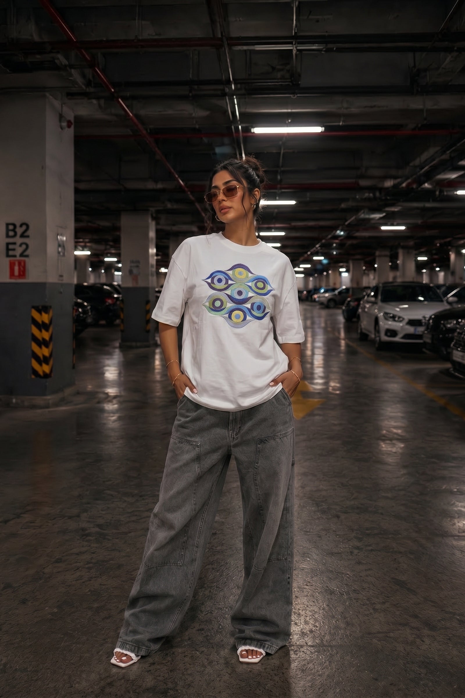 Person wearing a white t-shirt with a colorful embroidery design and gray pants in a parking garage.