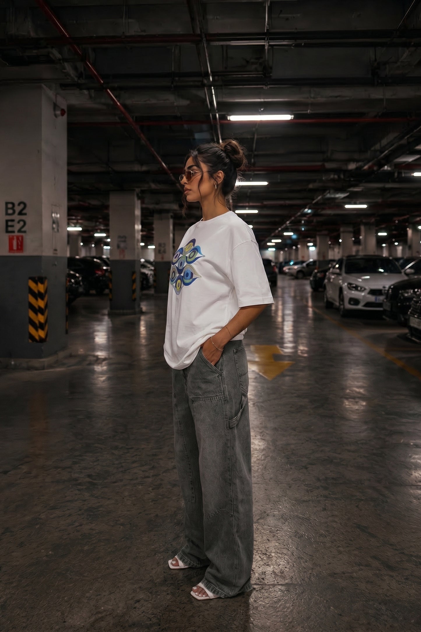 Person wearing a white t-shirt with a colorful embroidery design and gray pants in a parking garage.