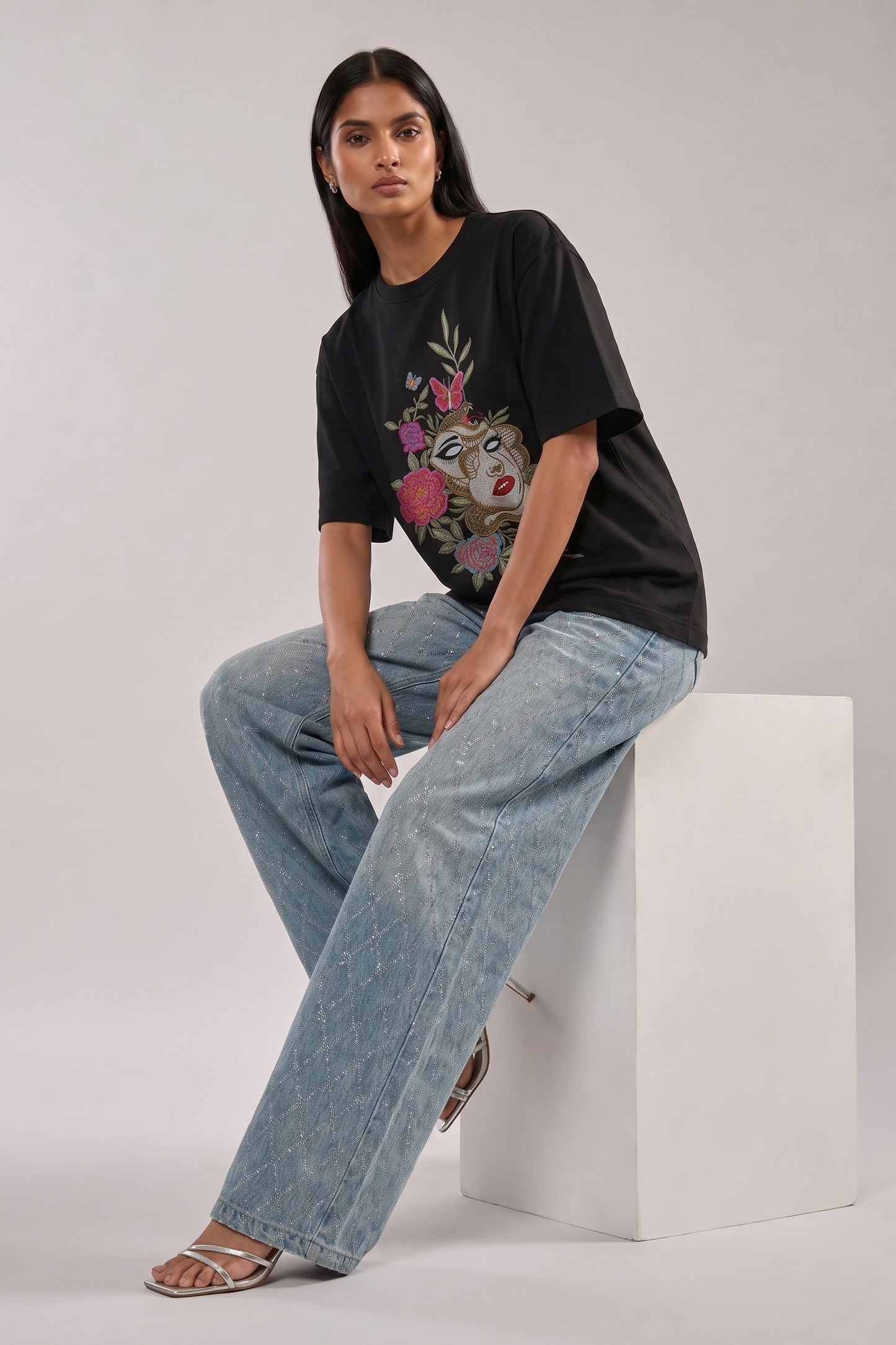 Woman wearing a black t-shirt with floral and butterfly embroidery design and blue jeans on a white background