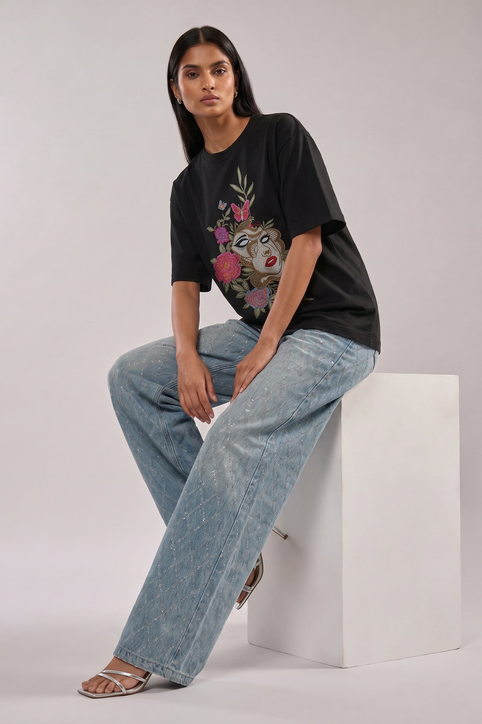 Woman wearing a black t-shirt with floral and butterfly embroidery design and blue jeans on a white background
