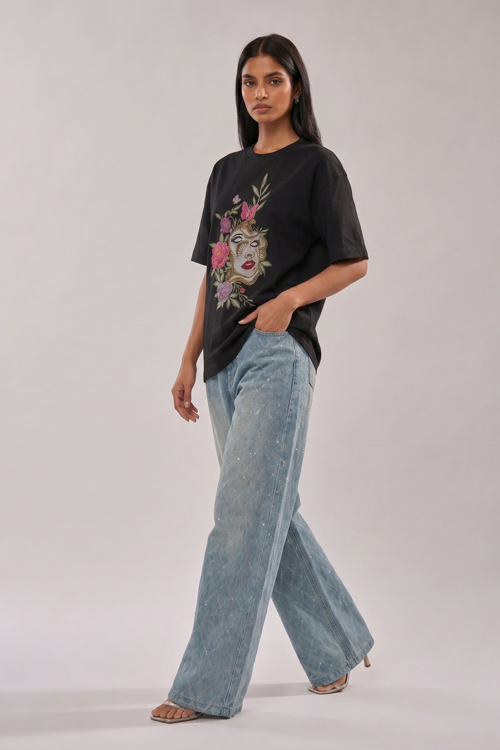 Woman wearing a black t-shirt with floral and butterfly embroidery design and blue jeans on a white background