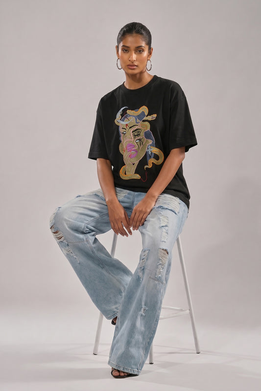 Person wearing a black t-shirt with a embroidery design and light blue jeans on a plain background