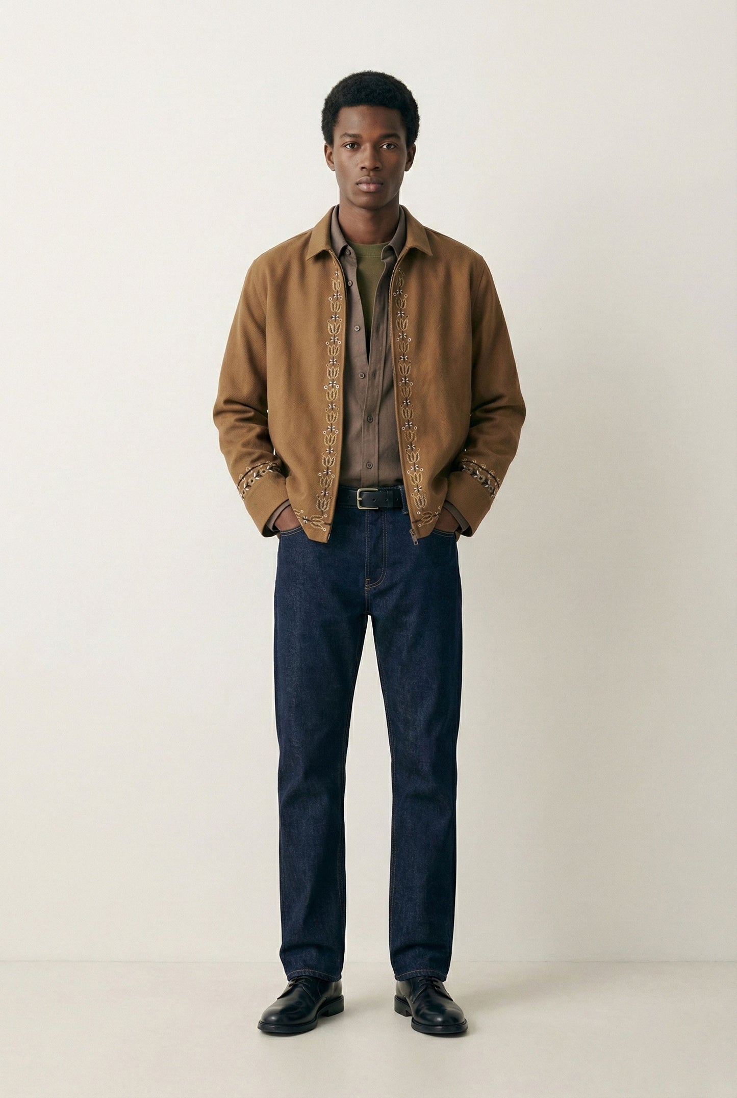 Man wearing a brown jacket, brown shirt, and dark blue jeans on a white background
