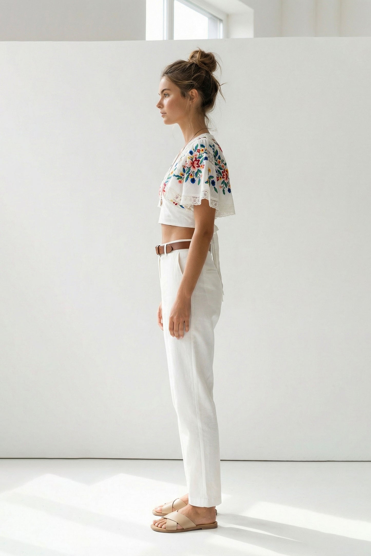 Woman wearing a floral top and white pants standing against a white wall.