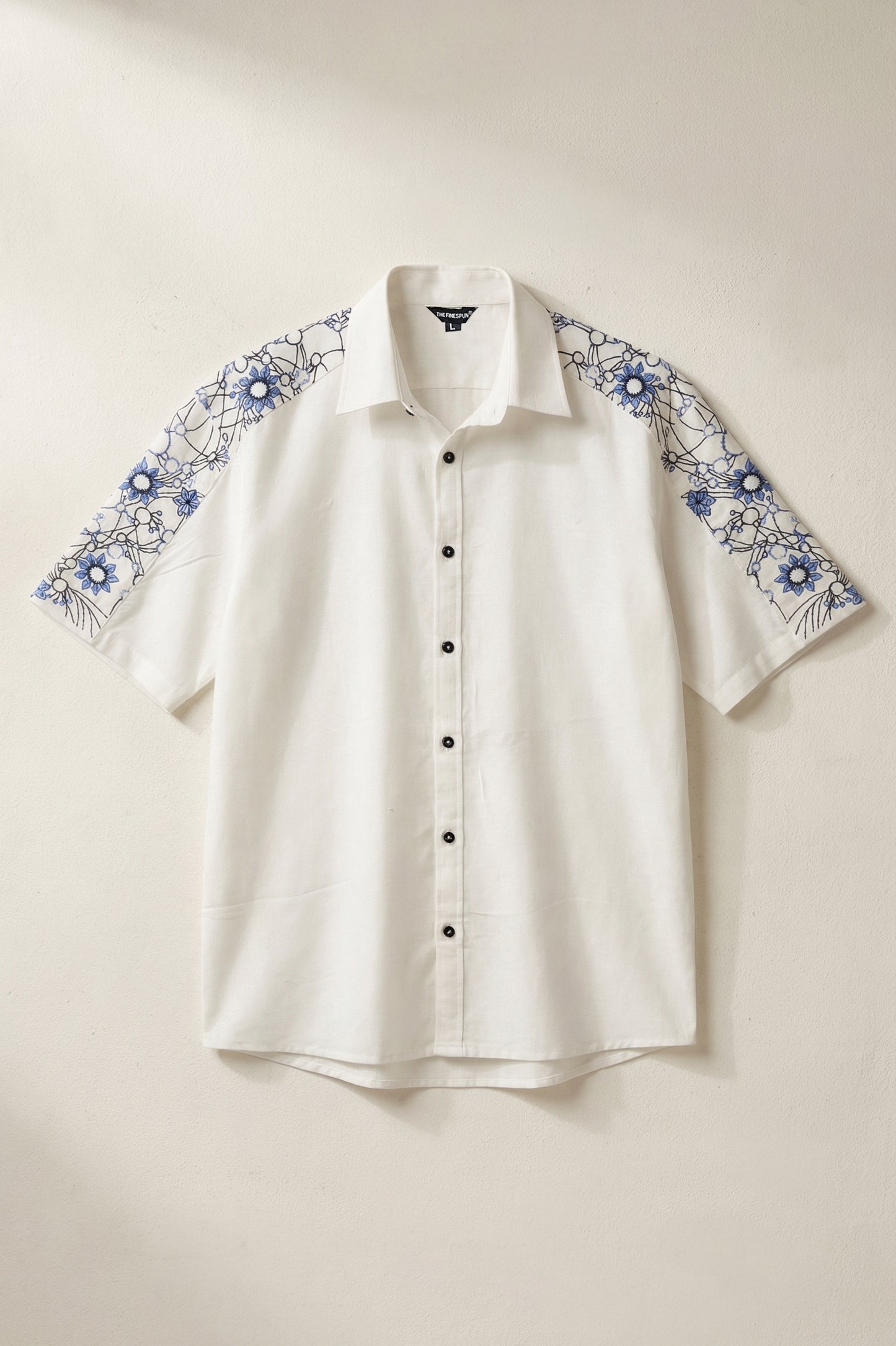 White shirt with blue floral embroidery on sleeves against a beige background