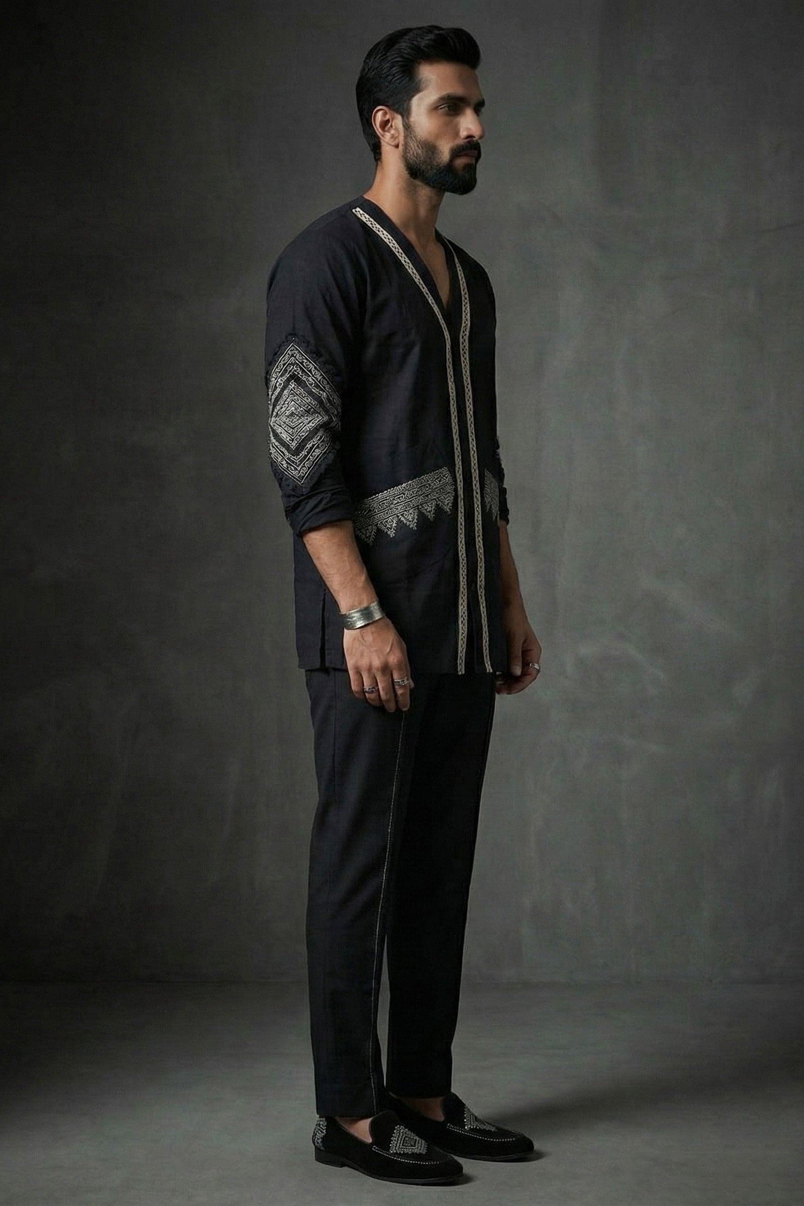 Man wearing a dark outfit with intricate patterns against a gray background