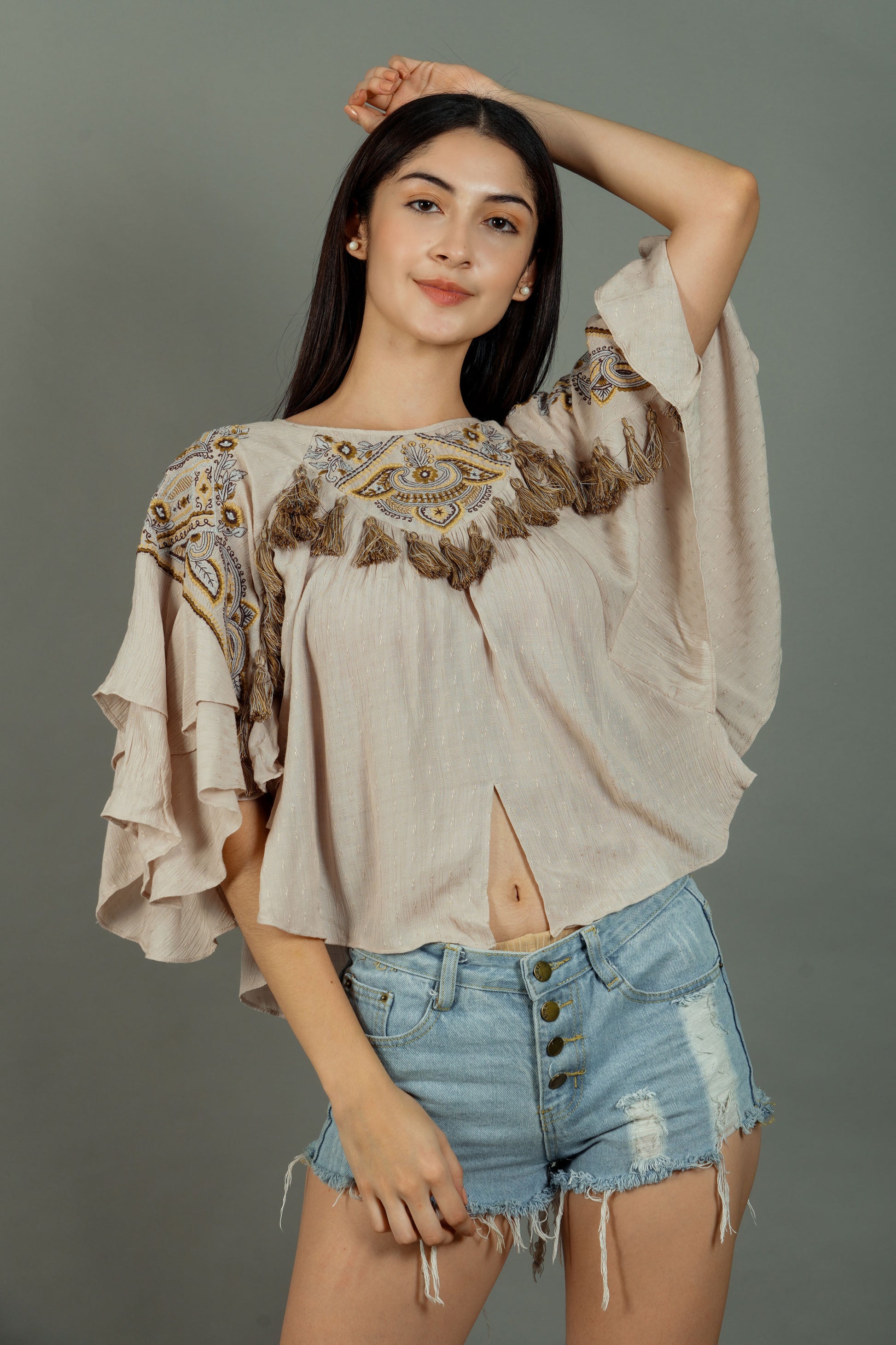 Woman wearing a beige blouse with intricate patterns and denim shorts on a gray background