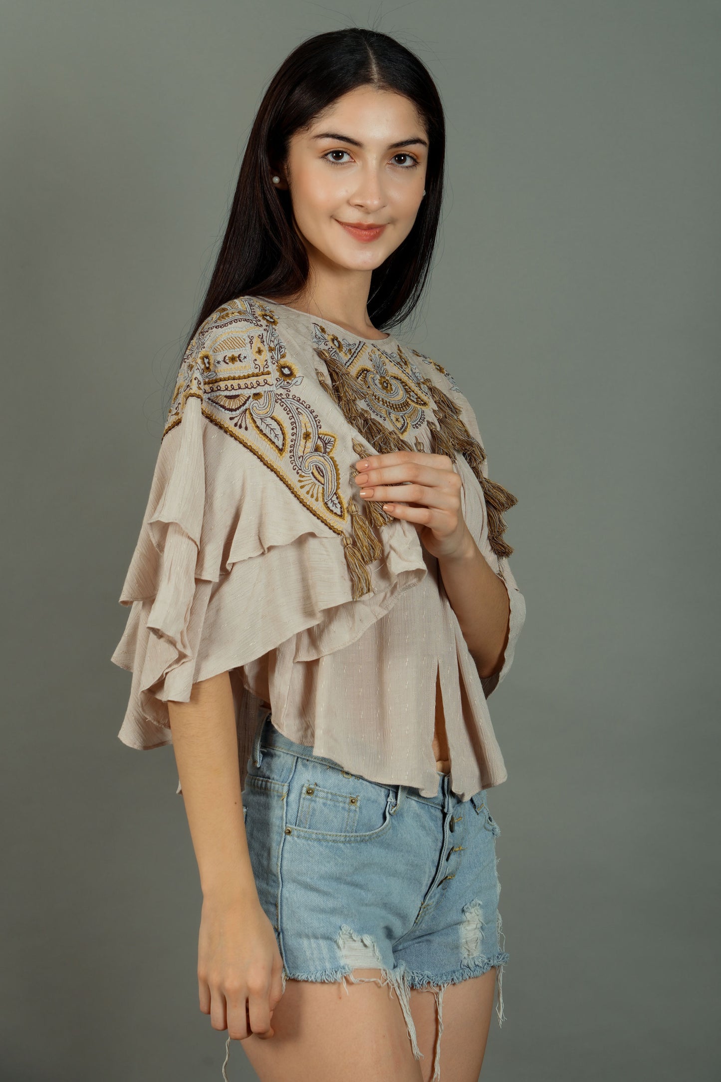 Woman wearing a beige embroidered top and denim shorts on a gray background