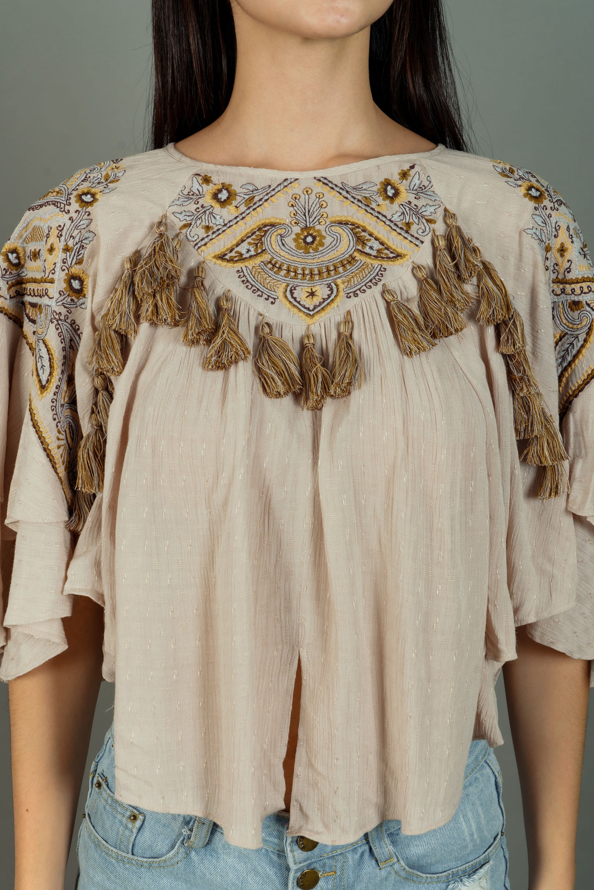 Beige blouse with intricate patterns and tassels worn by a model against a gray background