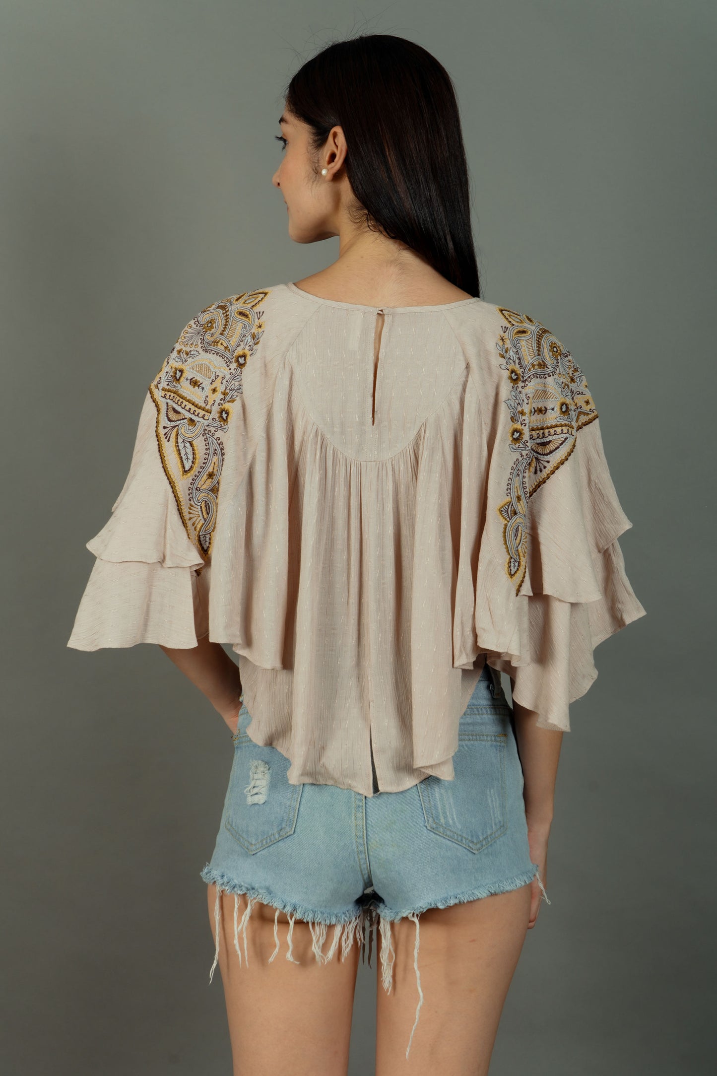 Woman wearing a beige blouse with intricate patterns and denim shorts on a gray background
