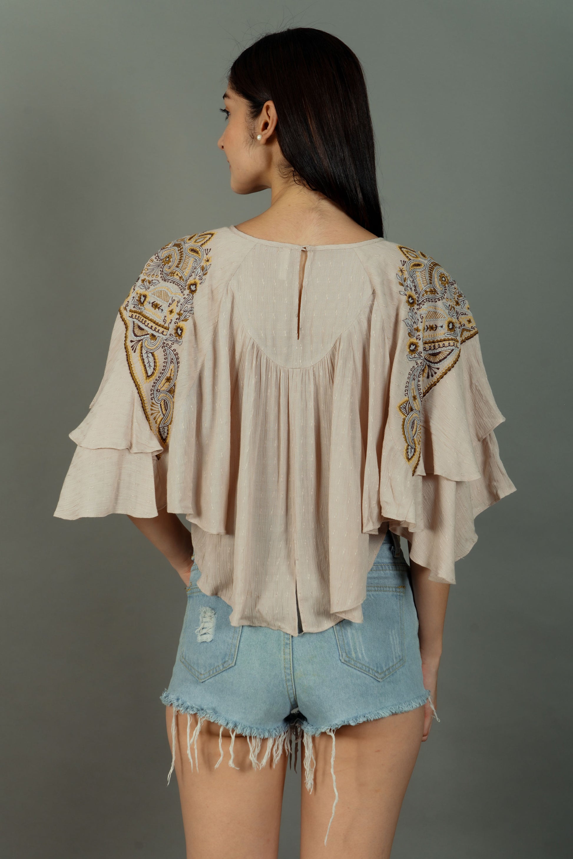 Woman wearing a beige blouse with intricate patterns and denim shorts on a gray background