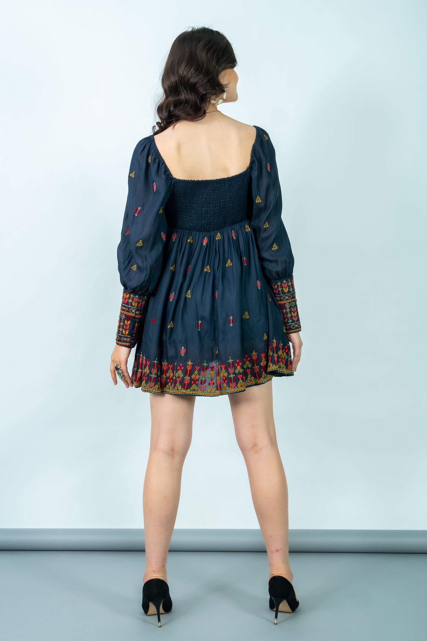 Woman wearing a dark blue dress with long sleeves and colorful patterns on a light gray background