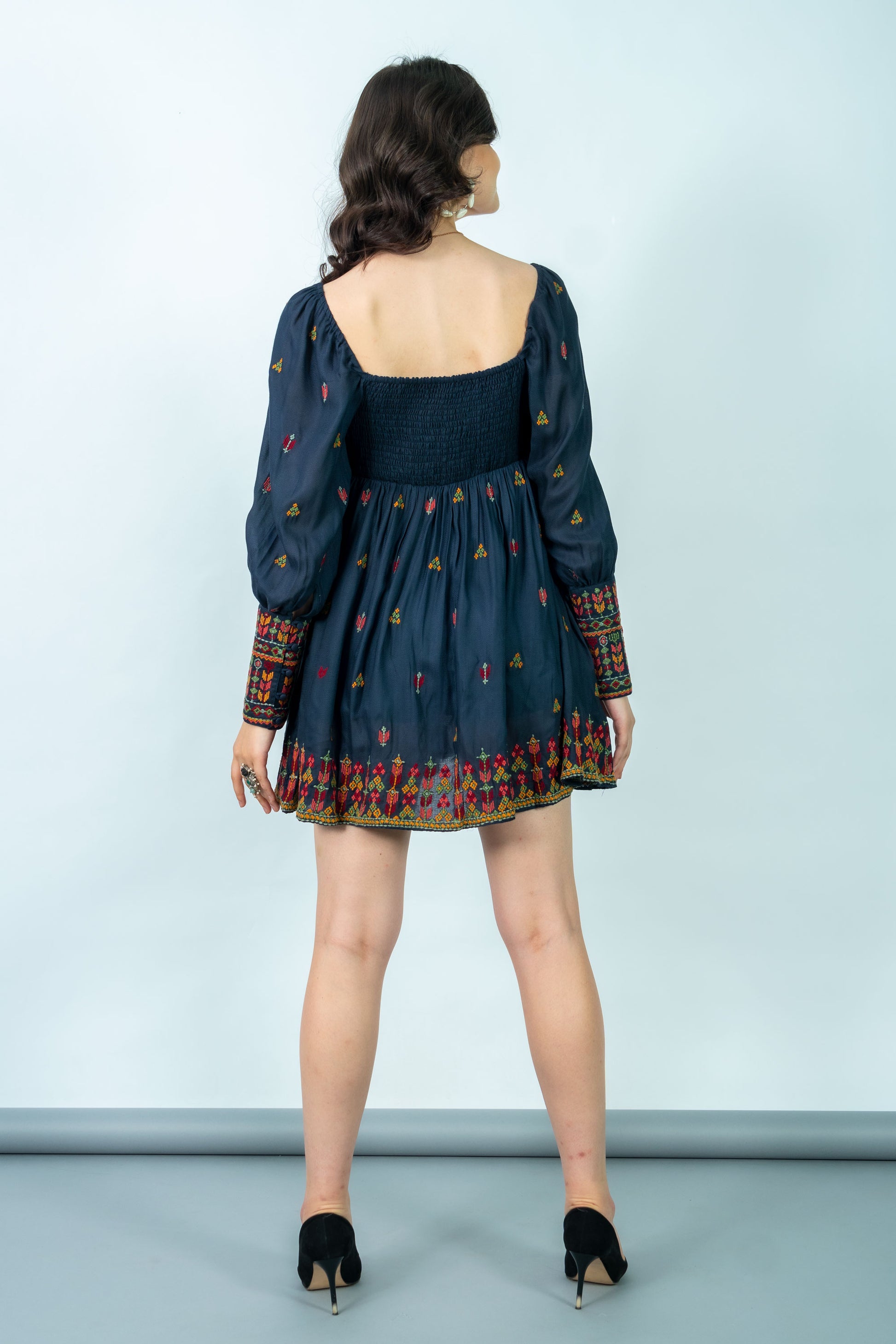 Woman wearing a dark blue dress with long sleeves and colorful patterns on a light gray background