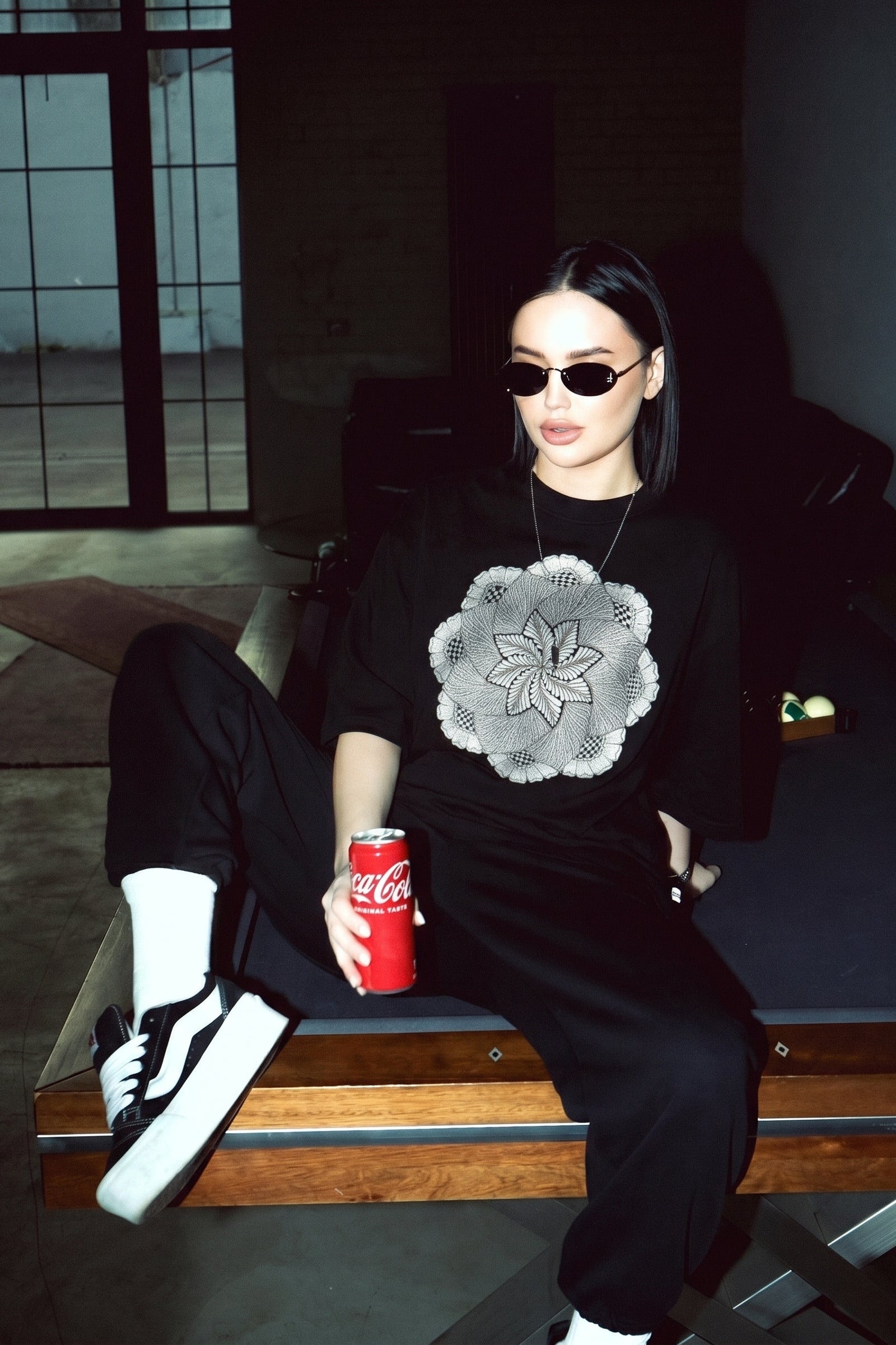Person wearing a black outfit with a white floral design, holding a Coca-Cola can in a dark room.