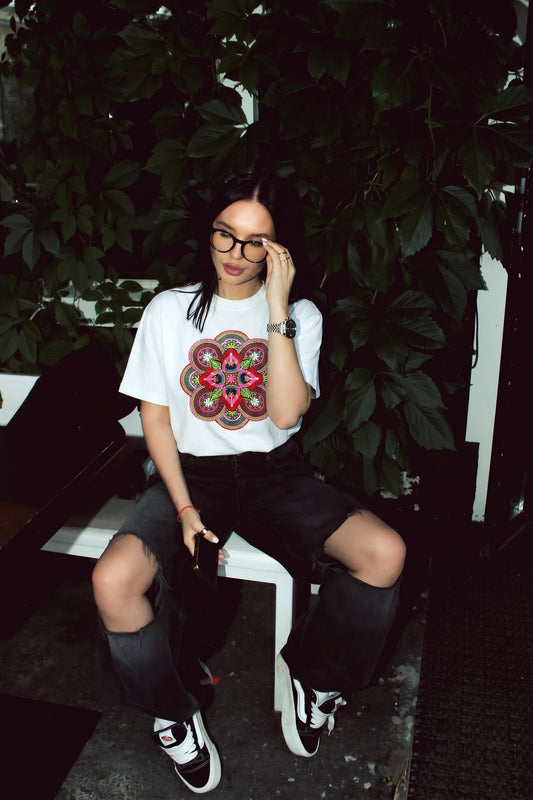 Woman sitting on a bench wearing a white t-shirt with a colorful design, black pants, and black shoes.