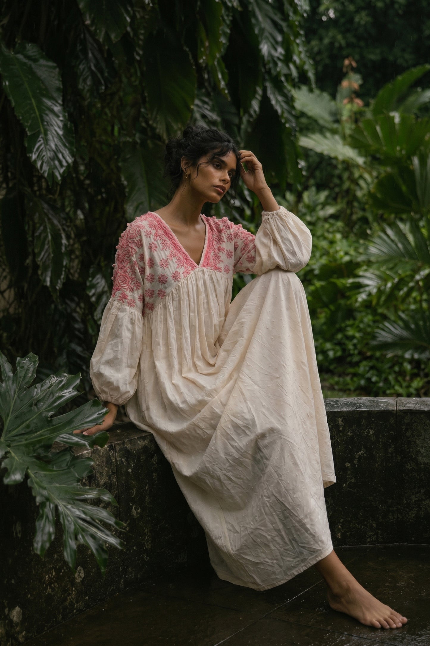 Woman in a long, light-colored dress with floral patterns sitting among green foliage.