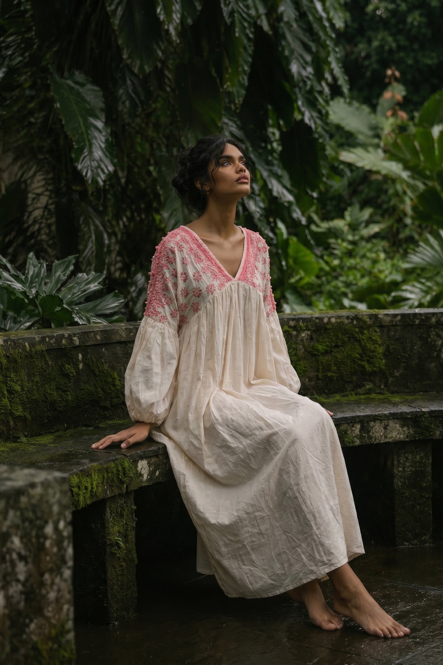 Woman in a white and pink dress sitting on a stone bench in a lush green forest.