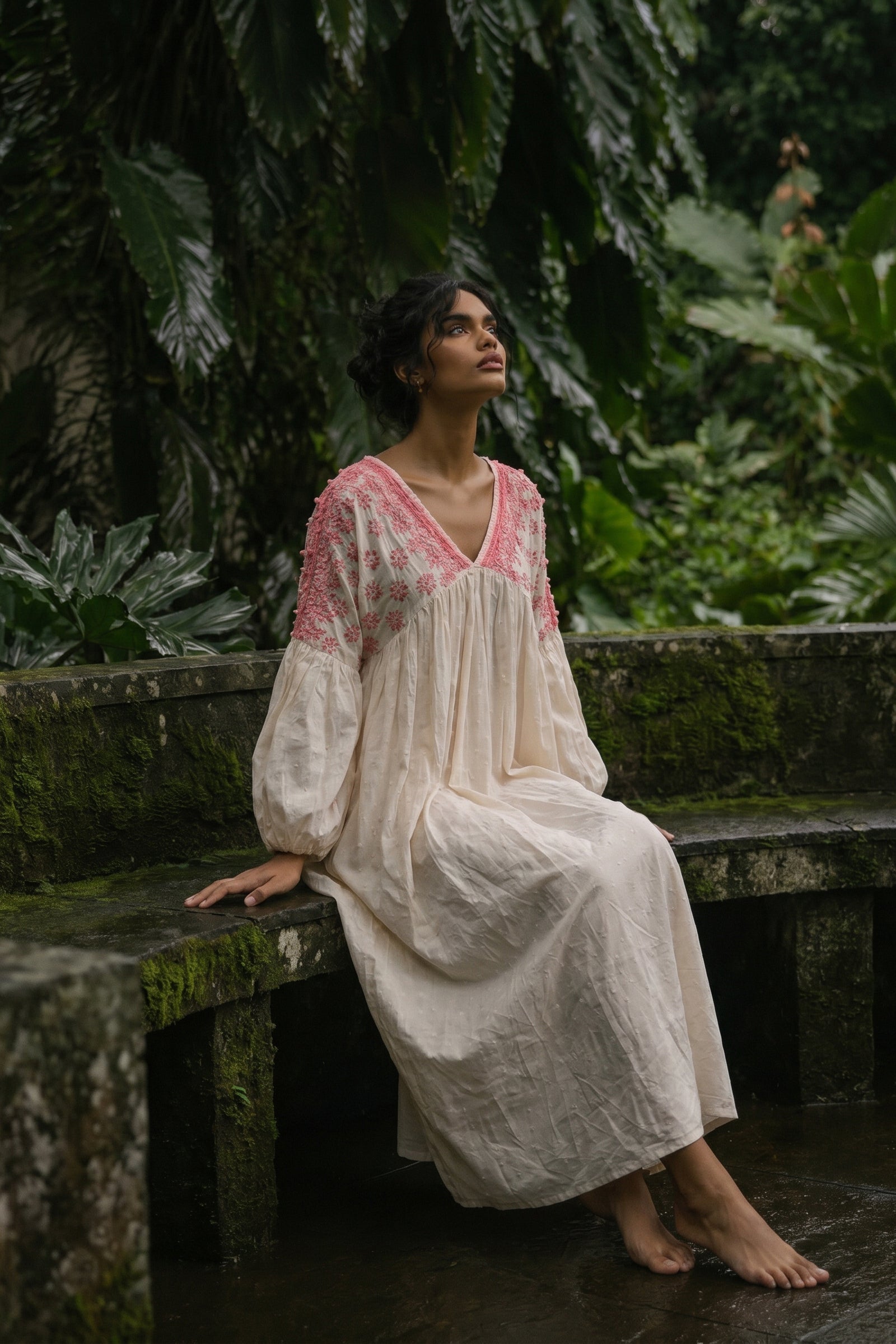 Woman in a white and pink dress sitting on a stone bench in a lush green forest.