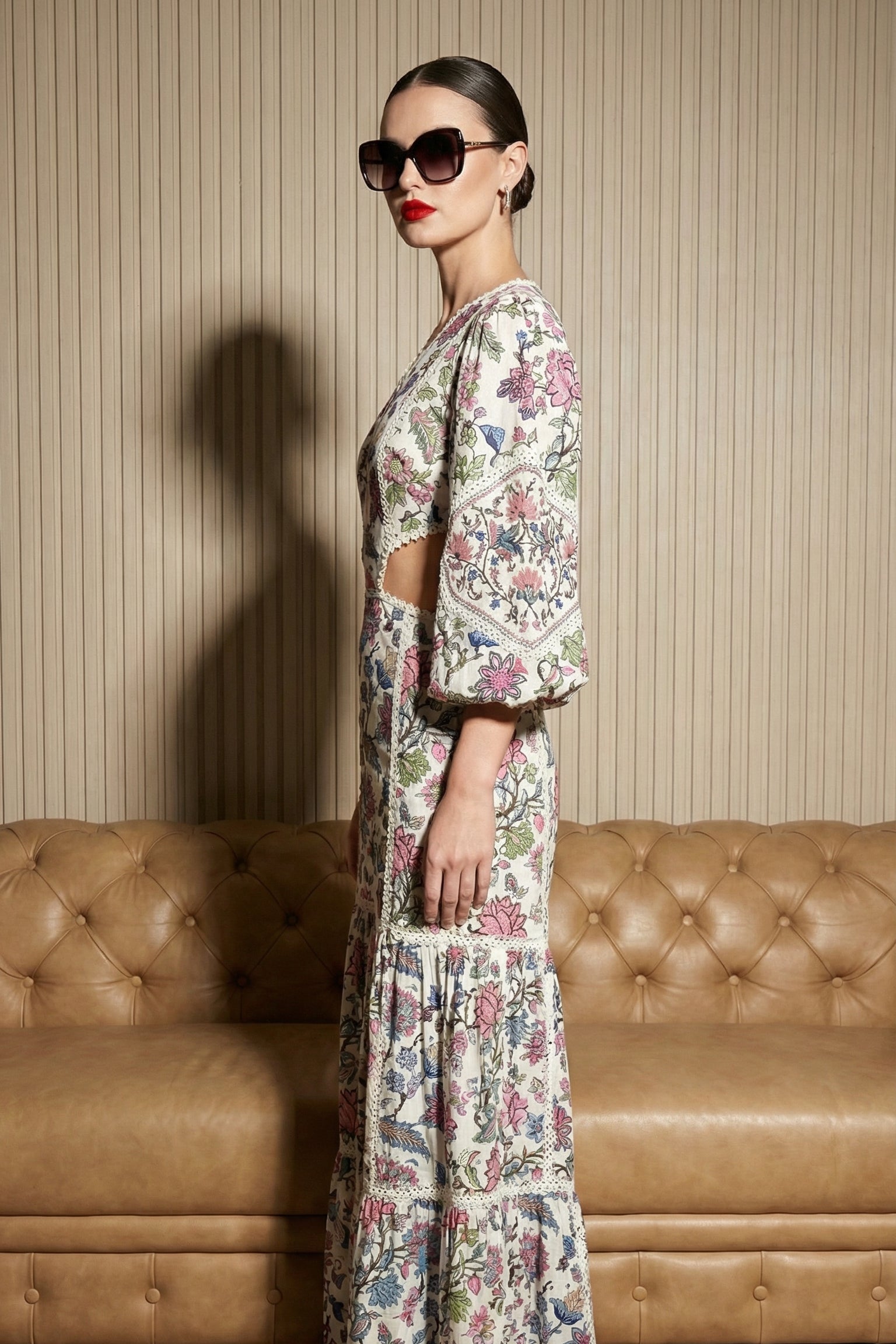 Woman in a floral outfit standing in front of a brown leather couch with a striped wall background.