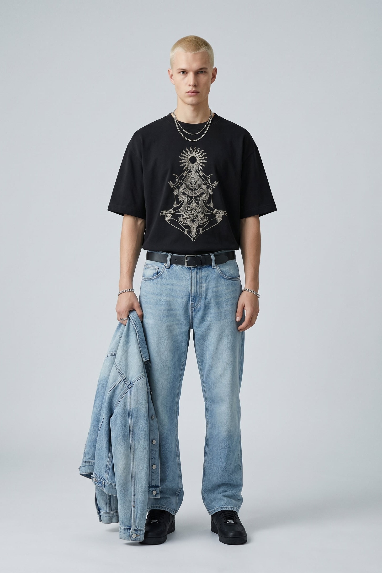Person wearing a black t-shirt with a graphic design, light blue jeans, and holding a denim jacket on a plain background