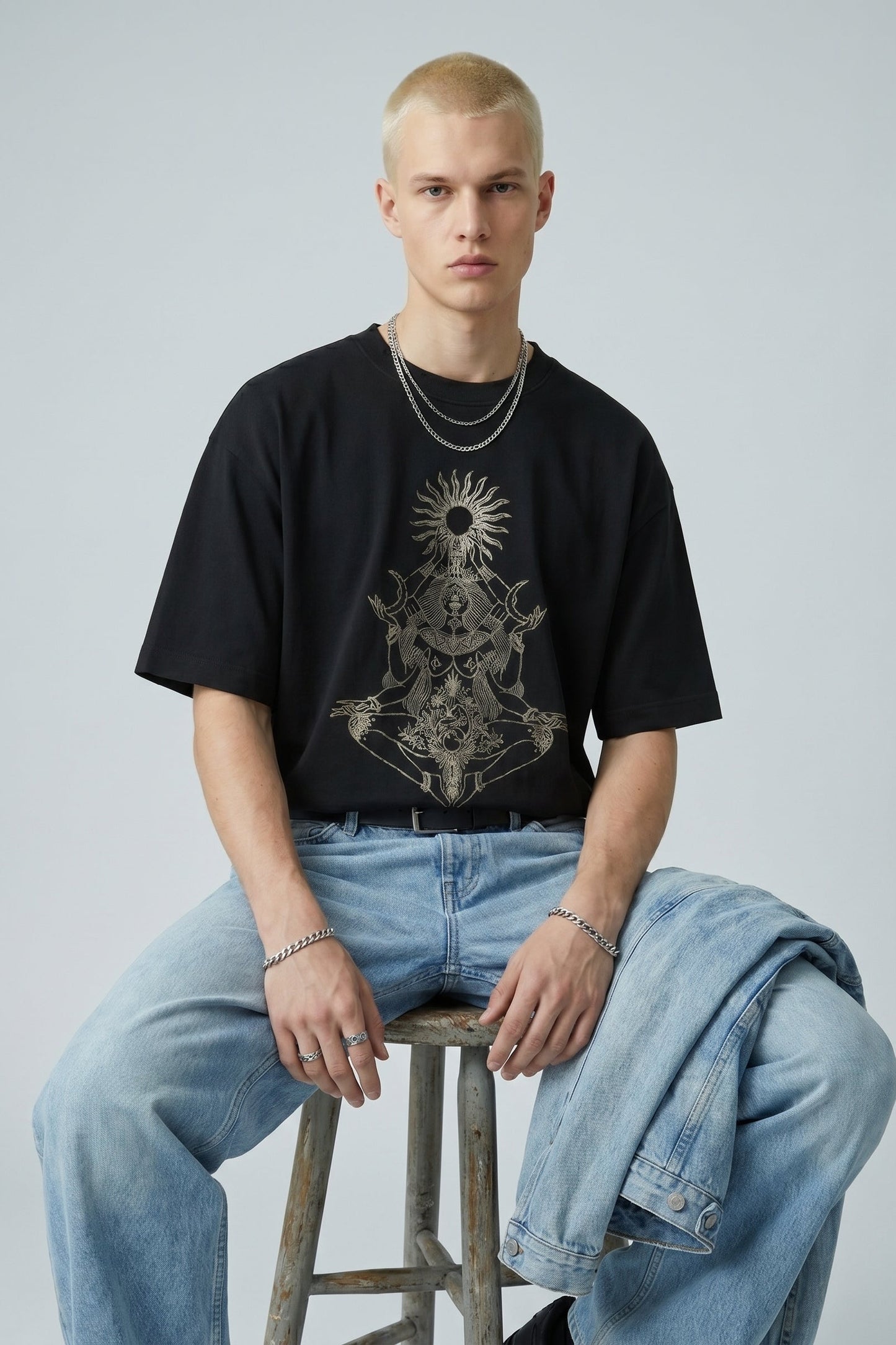Person wearing a black t-shirt with a graphic design and light blue jeans, sitting on a stool against a plain background.