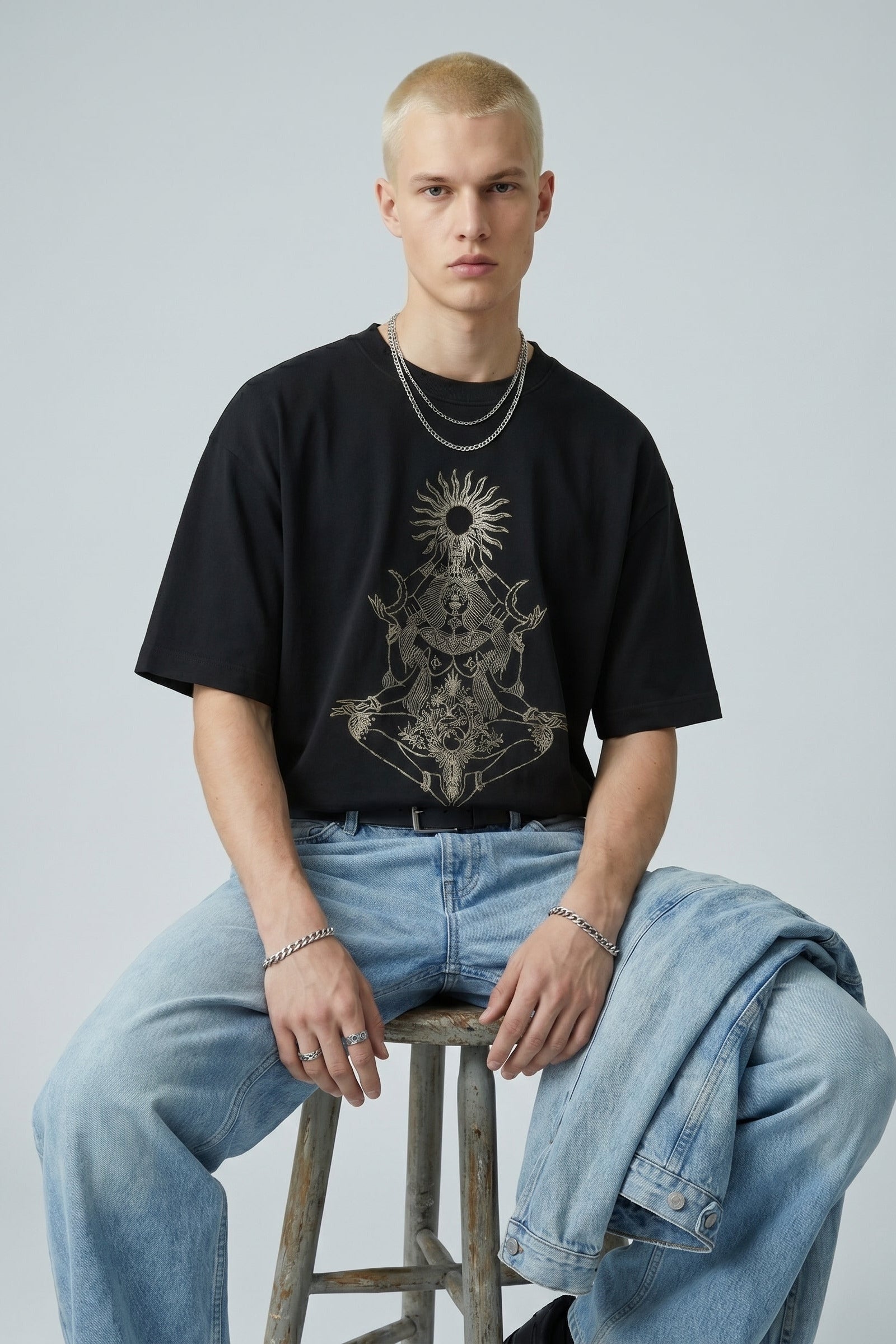 Person wearing a black t-shirt with a graphic design and light blue jeans, sitting on a stool against a plain background.