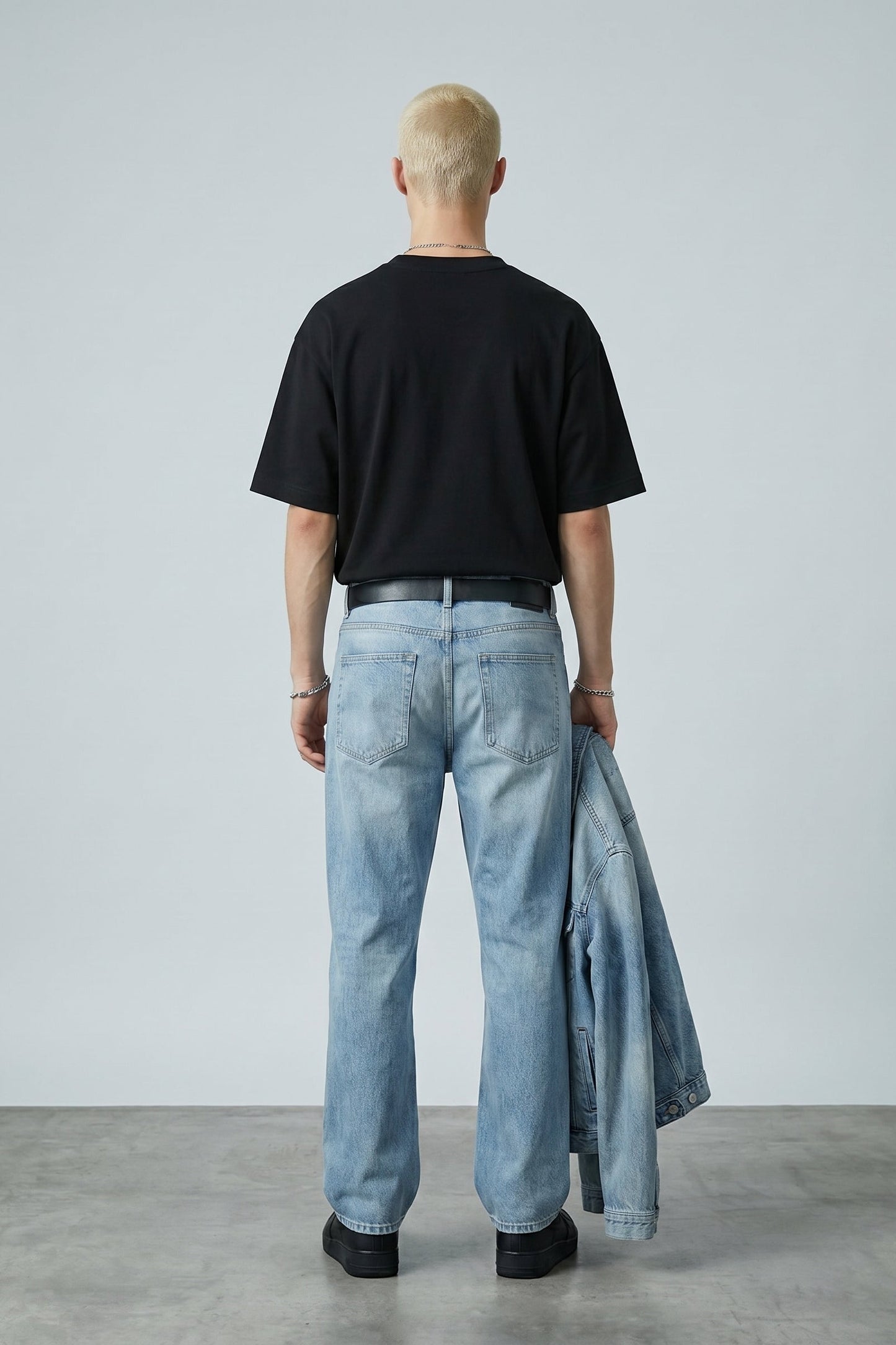 Person wearing a black t-shirt and light blue jeans holding a denim jacket against a plain background