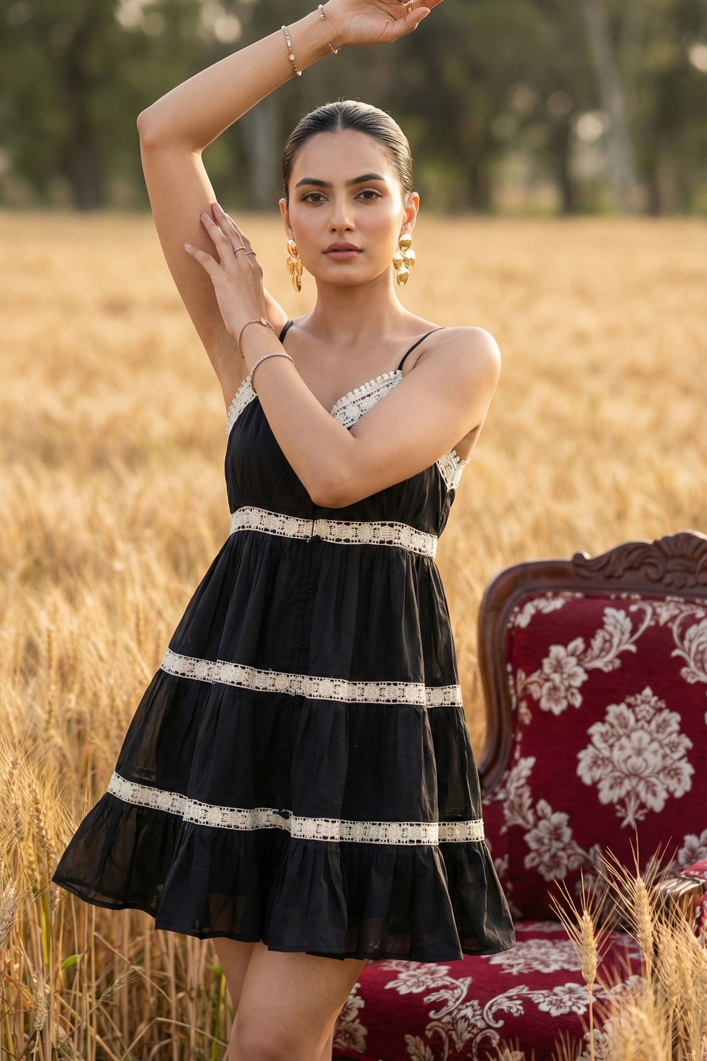 Woman in a black dress with white trim standing in a field with a chair and floral pattern in the background.