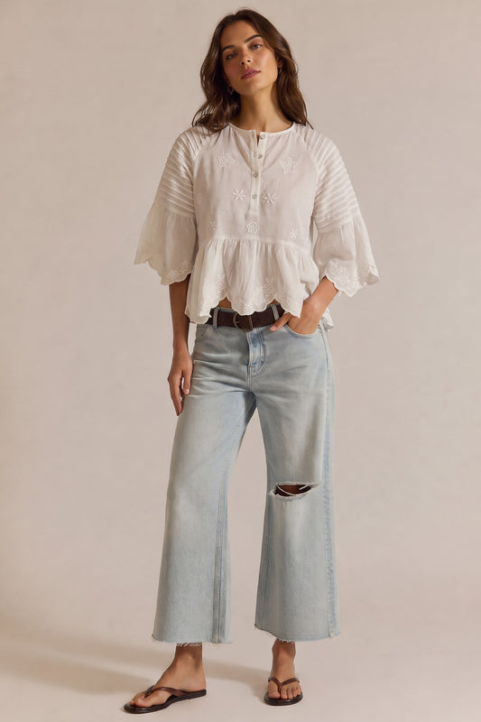 A woman modeling a white short-sleeve peplum blouse with gathered sleeves and light-wash distressed jeans, standing against a plain beige studio background.