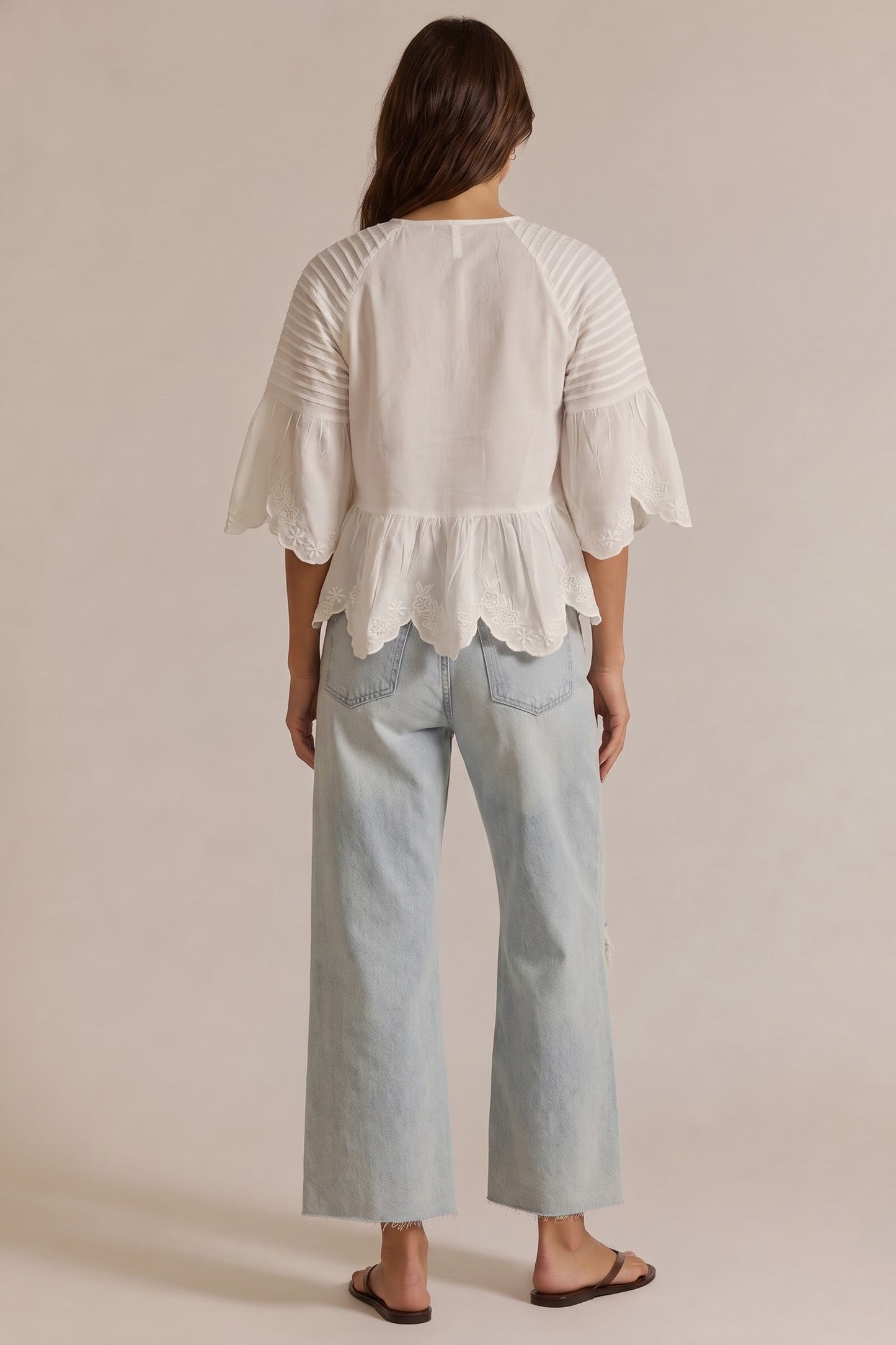 A woman viewed from the back wearing a white peplum blouse with ruffled sleeves and light-wash wide-leg jeans against a plain beige studio background.