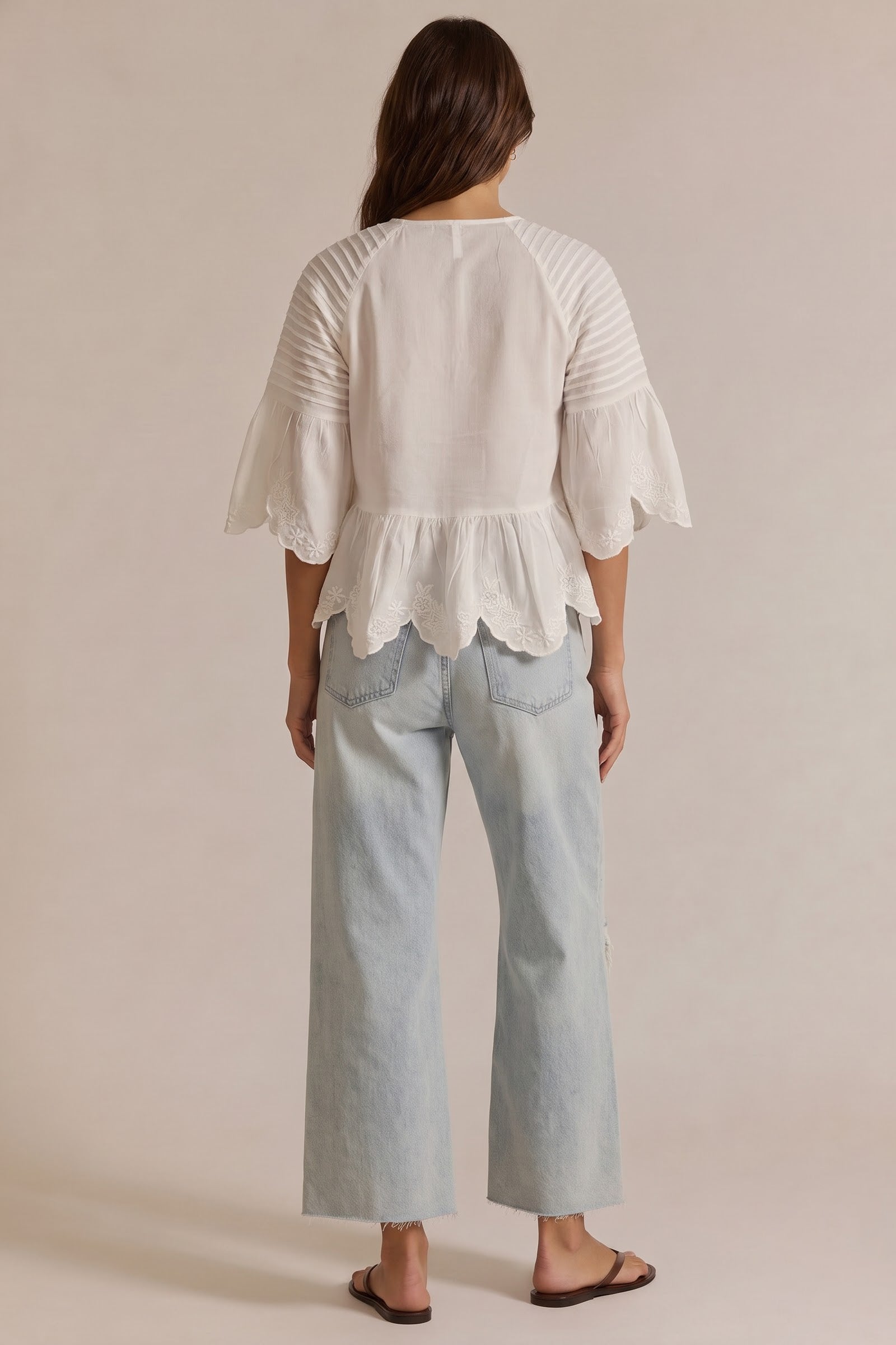 A woman viewed from the back wearing a white peplum blouse with ruffled sleeves and light-wash wide-leg jeans against a plain beige studio background.