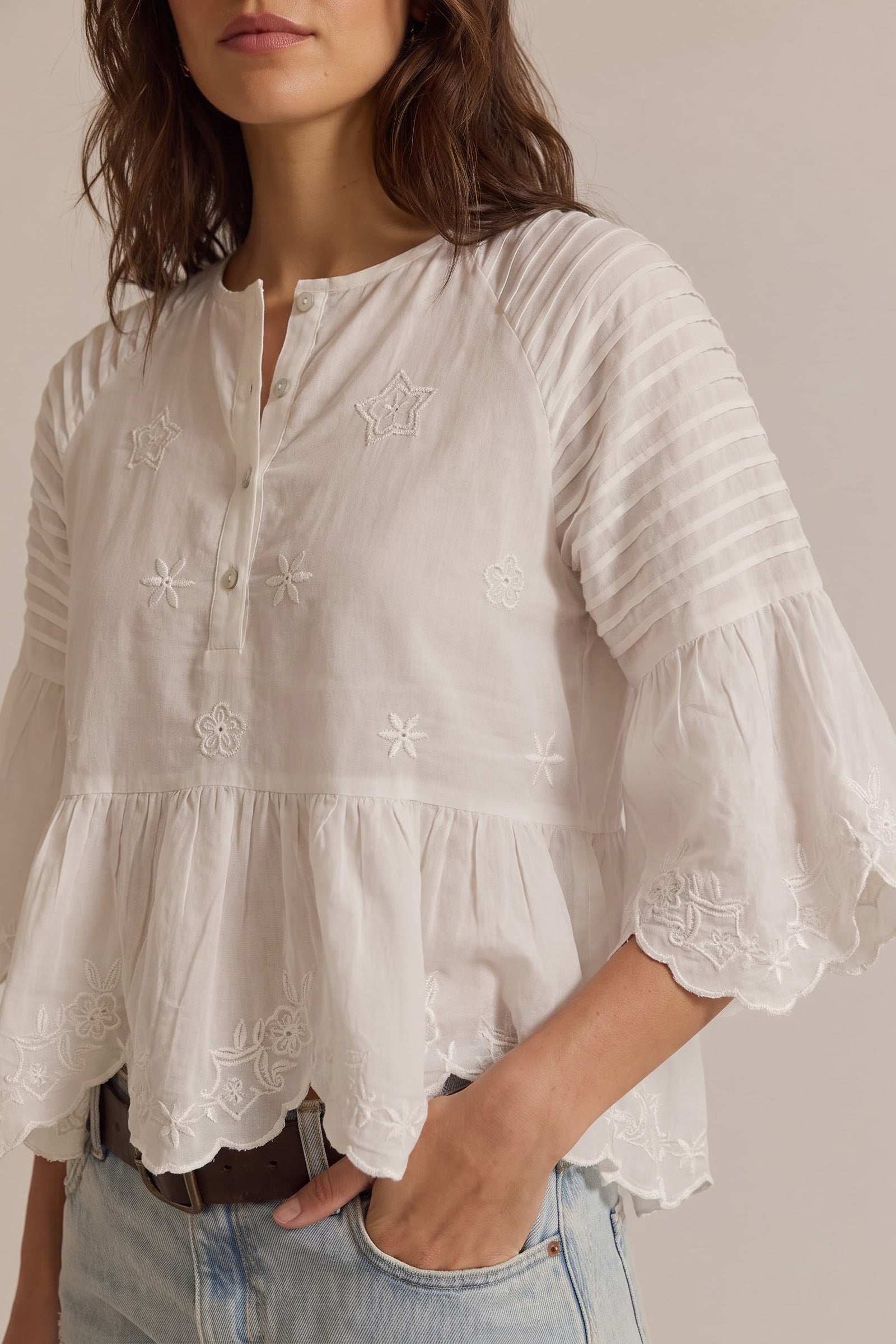 A woman wearing a white long-sleeve blouse with floral embroidery, a buttoned split neckline, and ruffled bell sleeves, paired with light-wash jeans against a plain beige background.