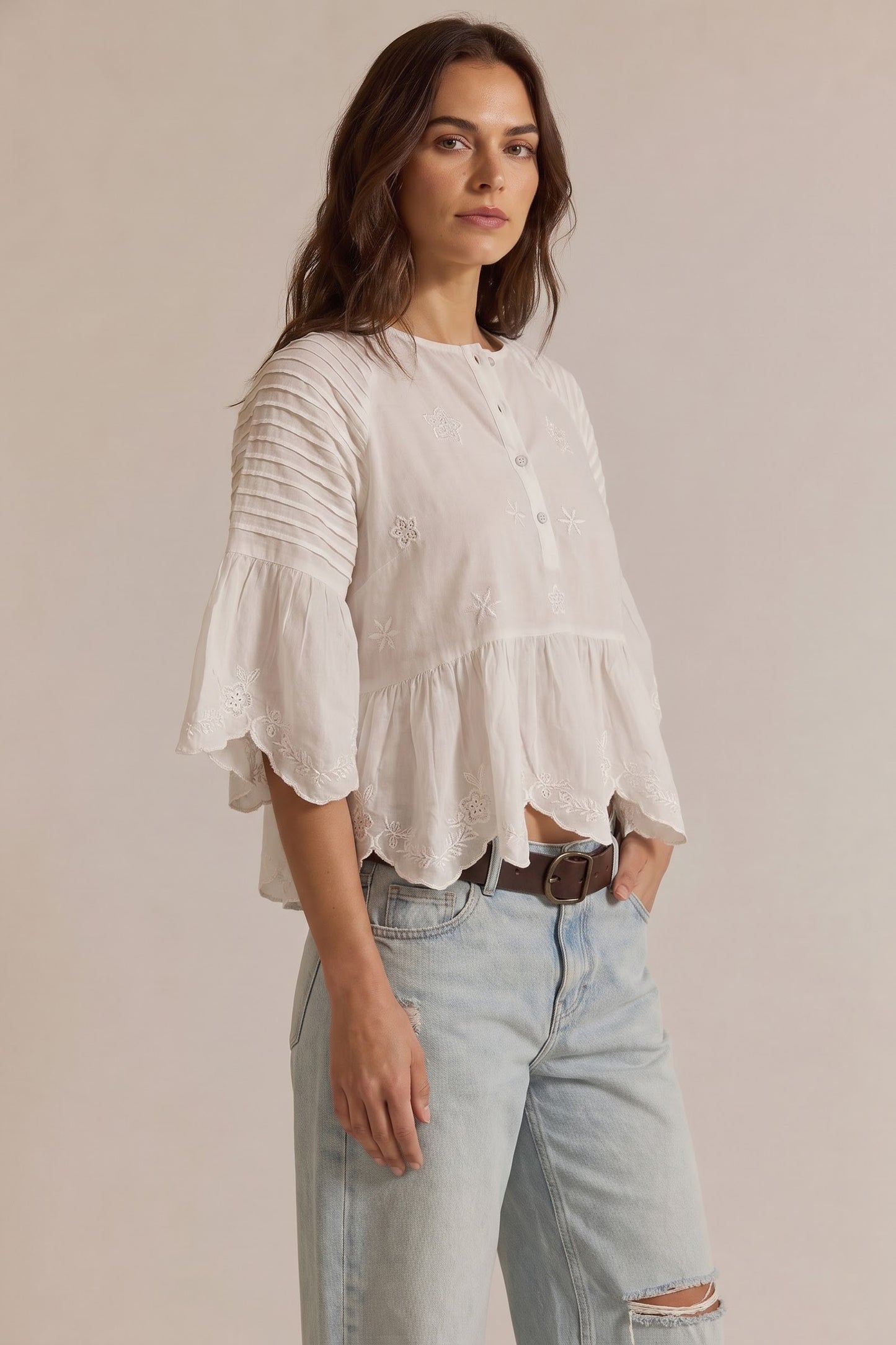 A woman modeling a white bohemian-style blouse with pleated sleeves, a buttoned collar, and a ruffled hem, paired with light-wash jeans against a neutral beige background.