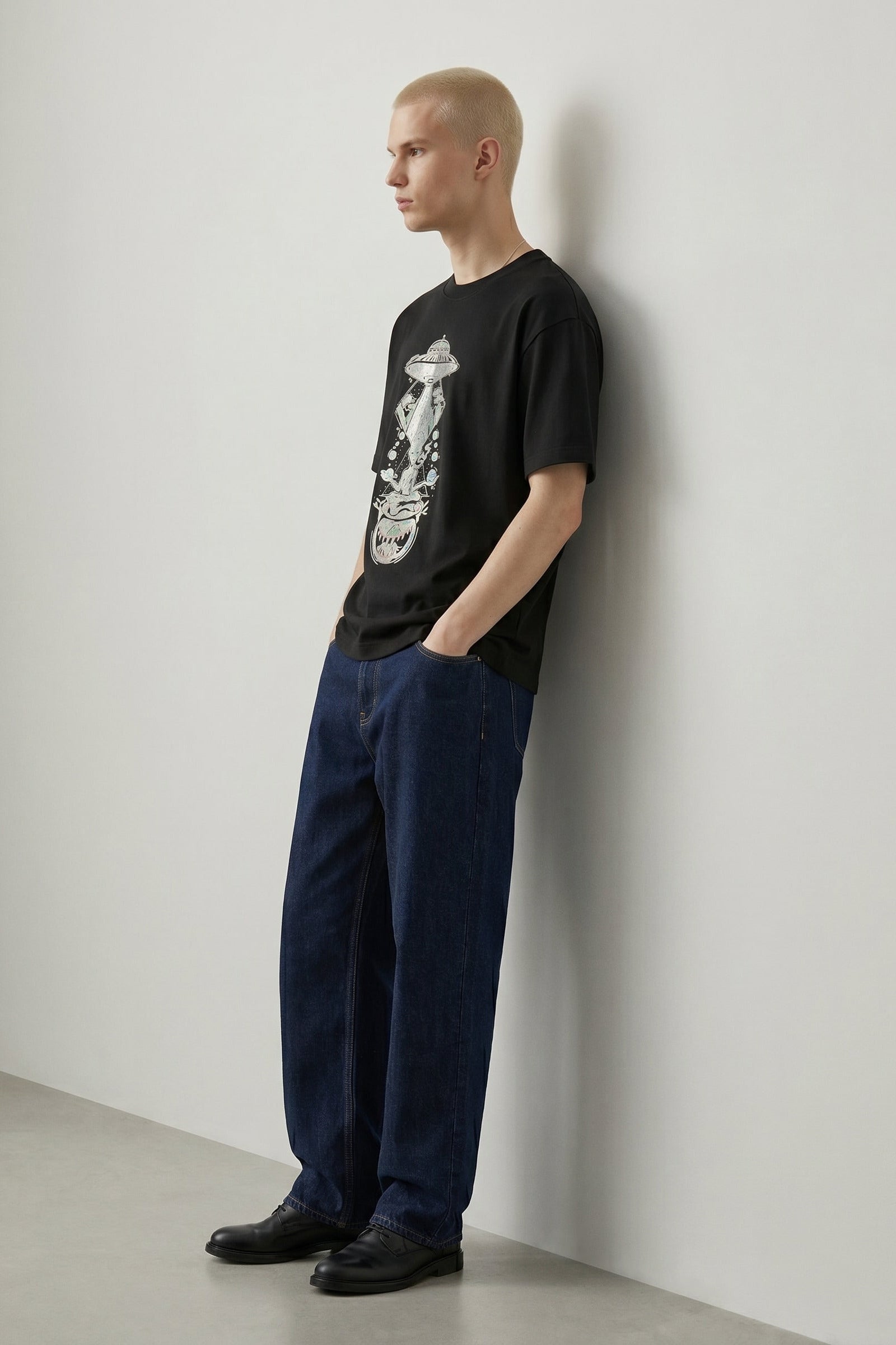 Person wearing a black t-shirt with a graphic design and blue pants against a white wall.