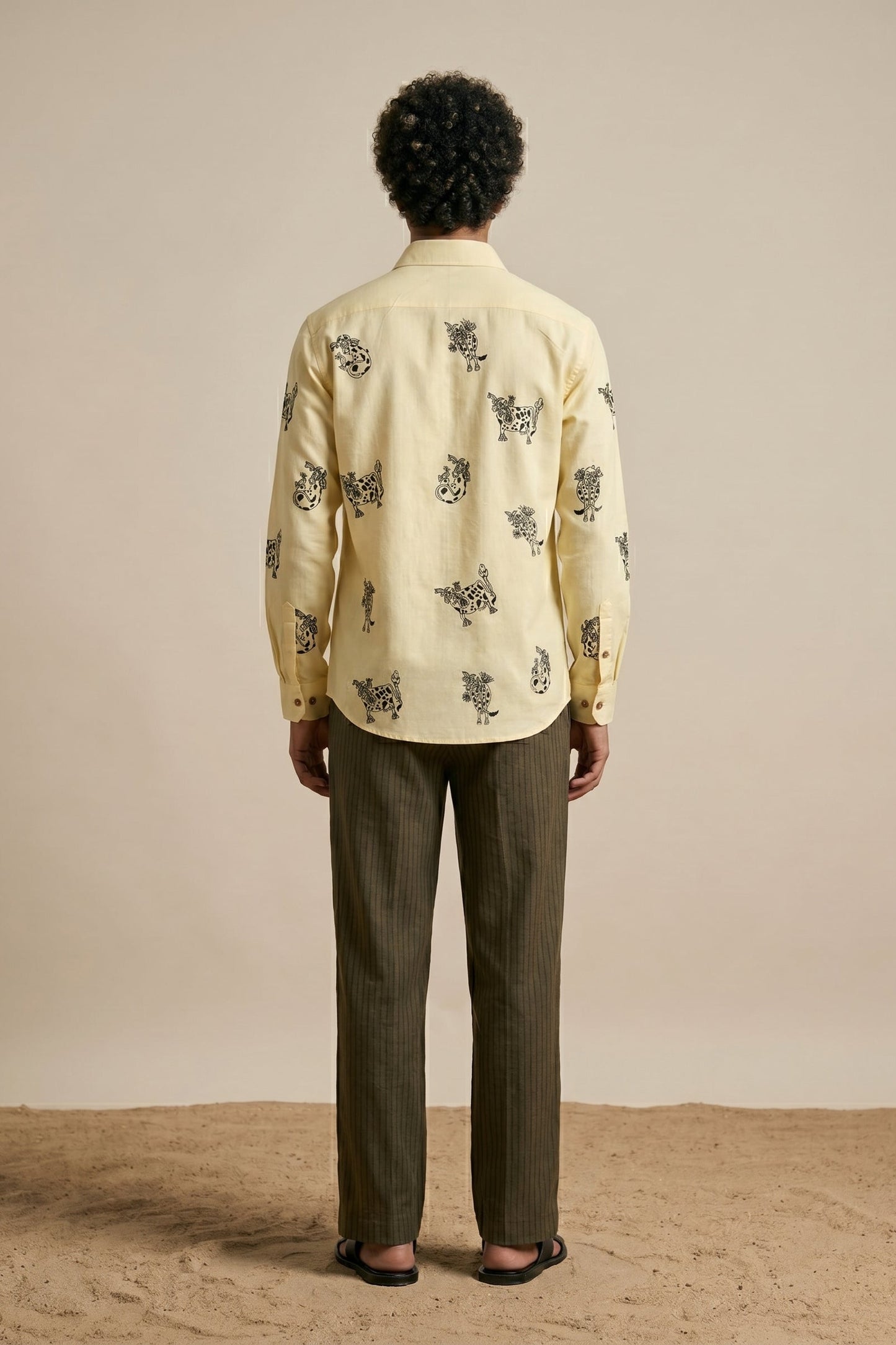Person wearing a light-colored shirt with a pattern and dark pants against a beige background