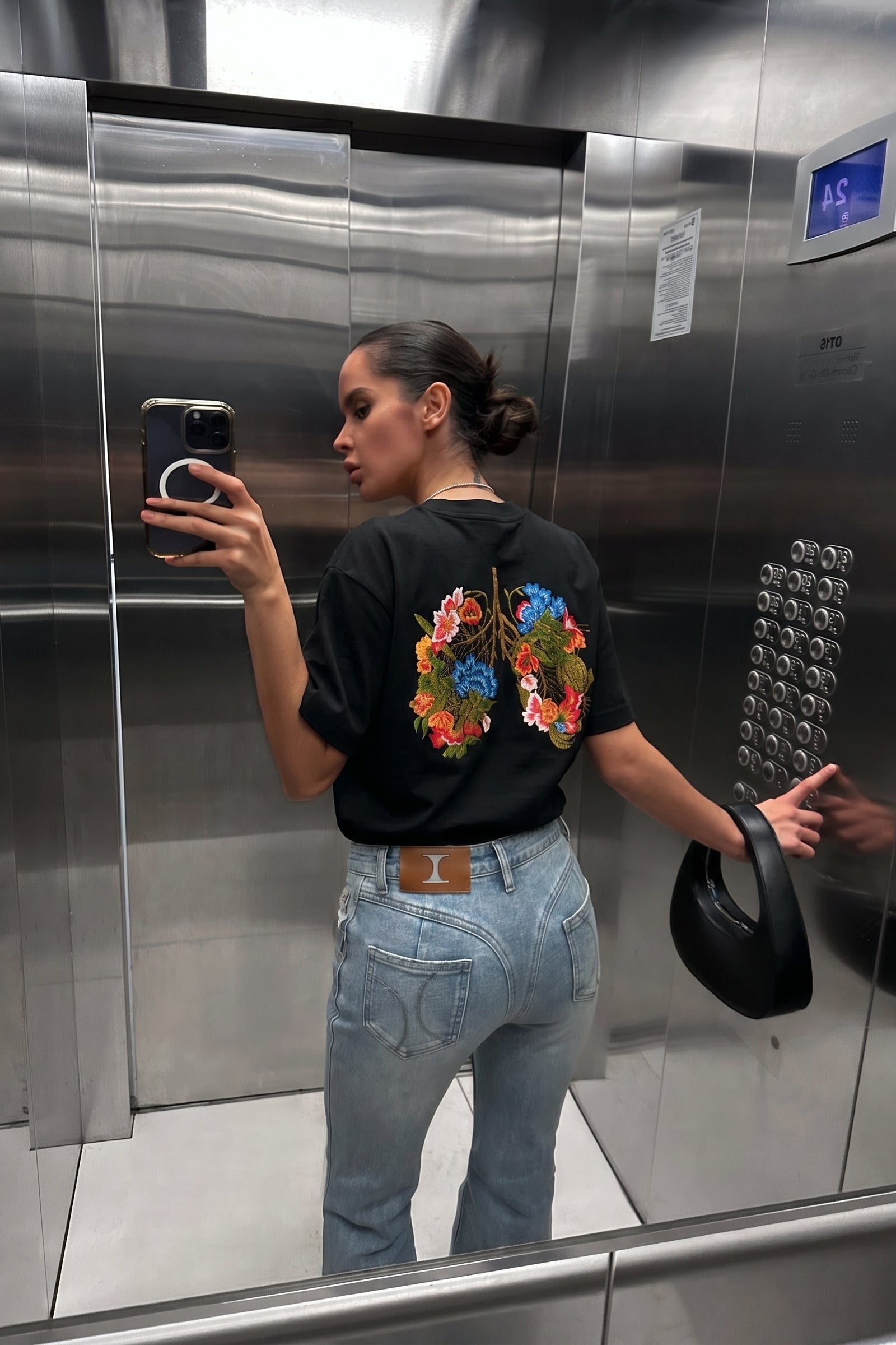 Person taking a mirror selfie in an elevator wearing a black t-shirt with colorful floral design.