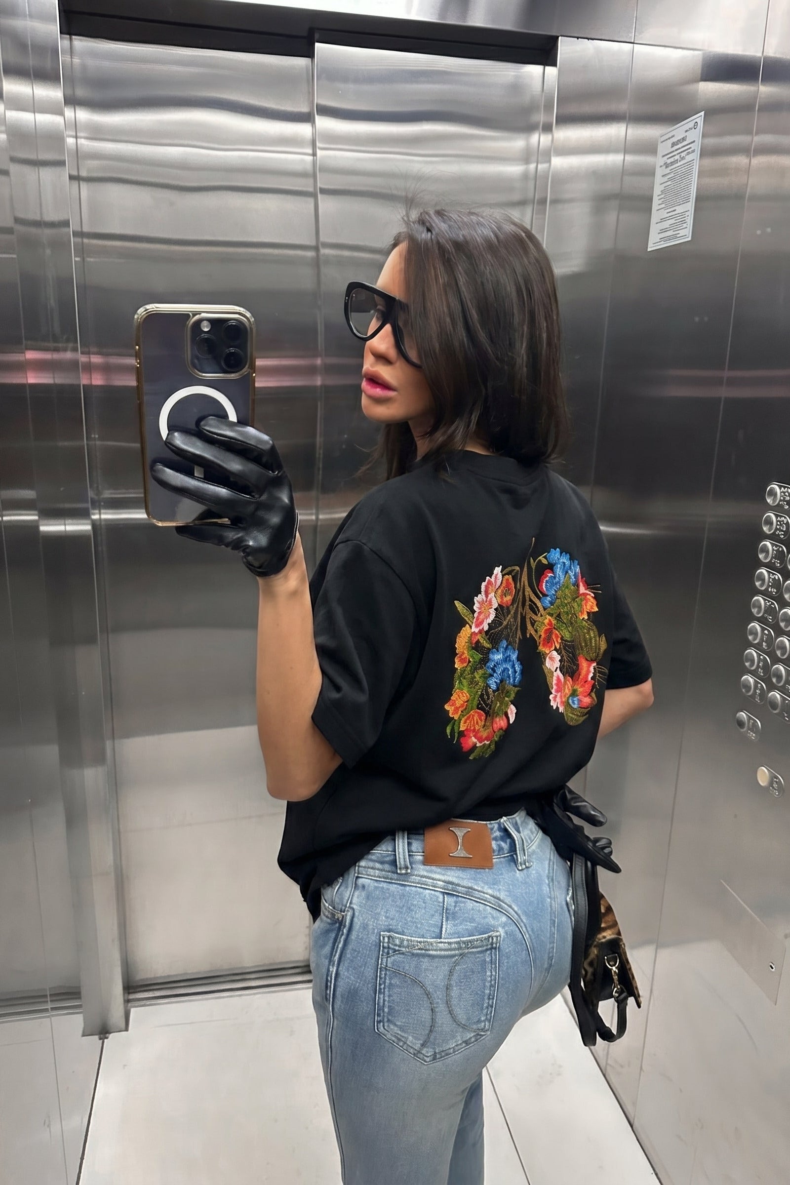 Person taking a mirror selfie in an elevator wearing a black t-shirt with colorful designs and light blue jeans.