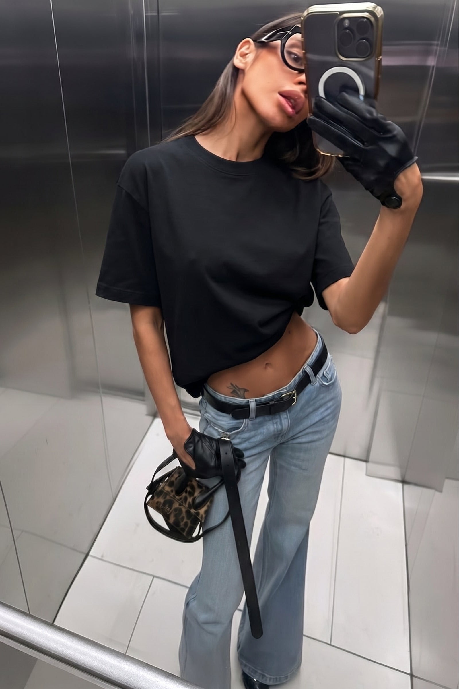 Person taking a mirror selfie wearing a black t-shirt, blue jeans, and black gloves in an elevator.