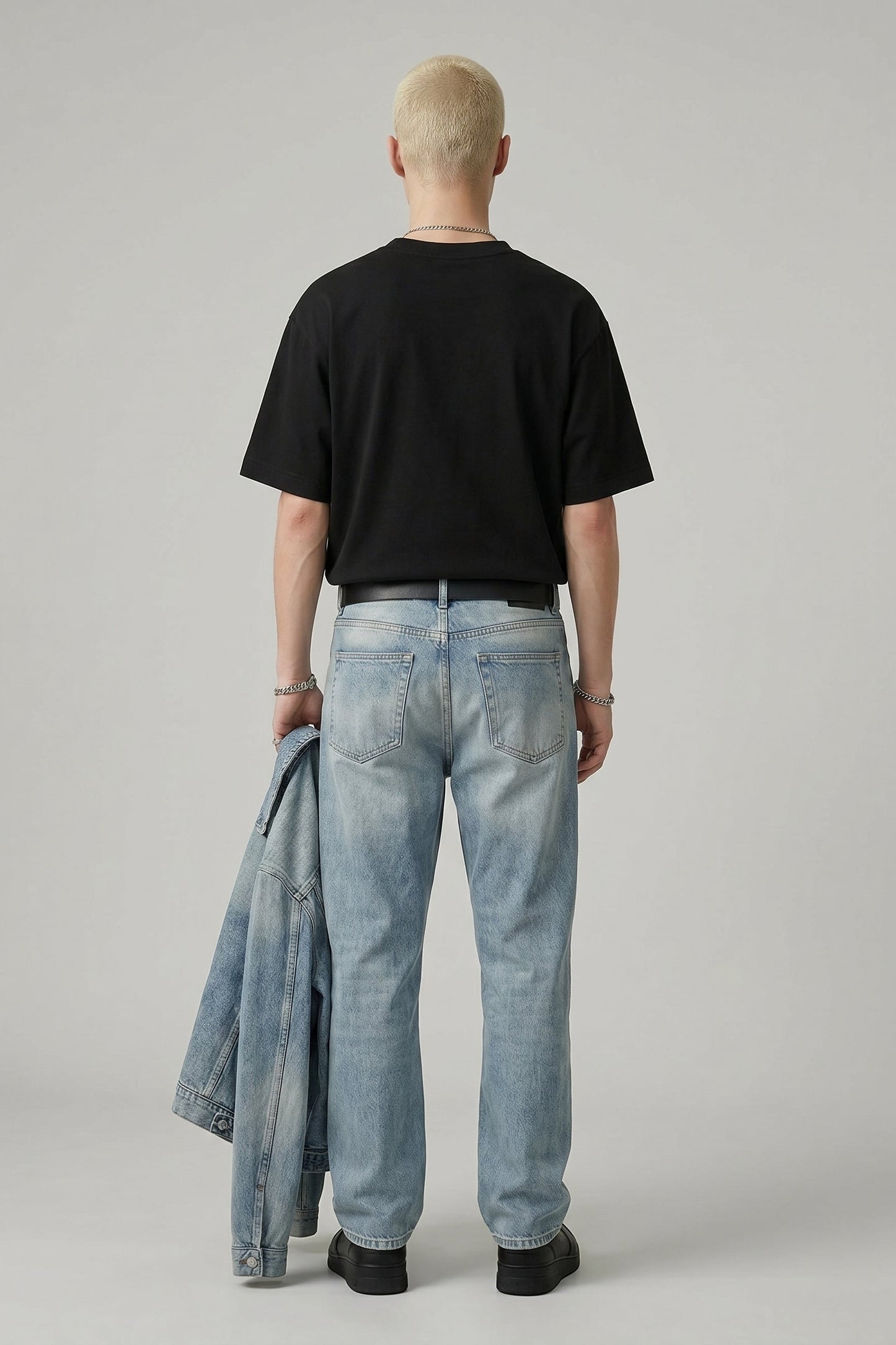 Person wearing a black t-shirt and light blue jeans holding a denim jacket on a plain background