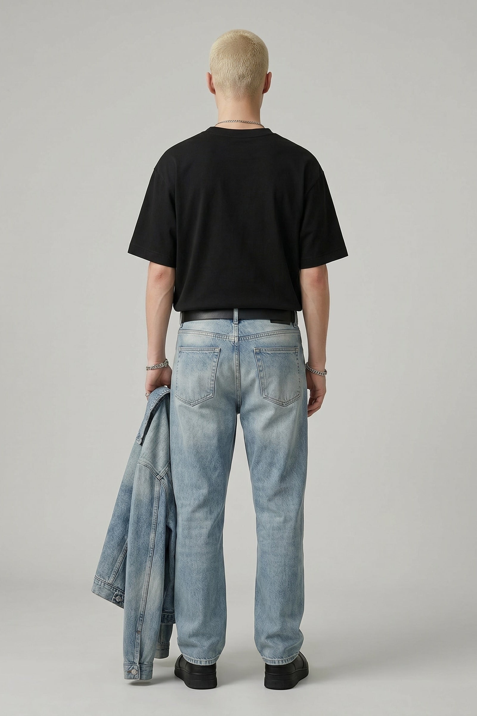 Person wearing a black t-shirt and light blue jeans holding a denim jacket on a plain background
