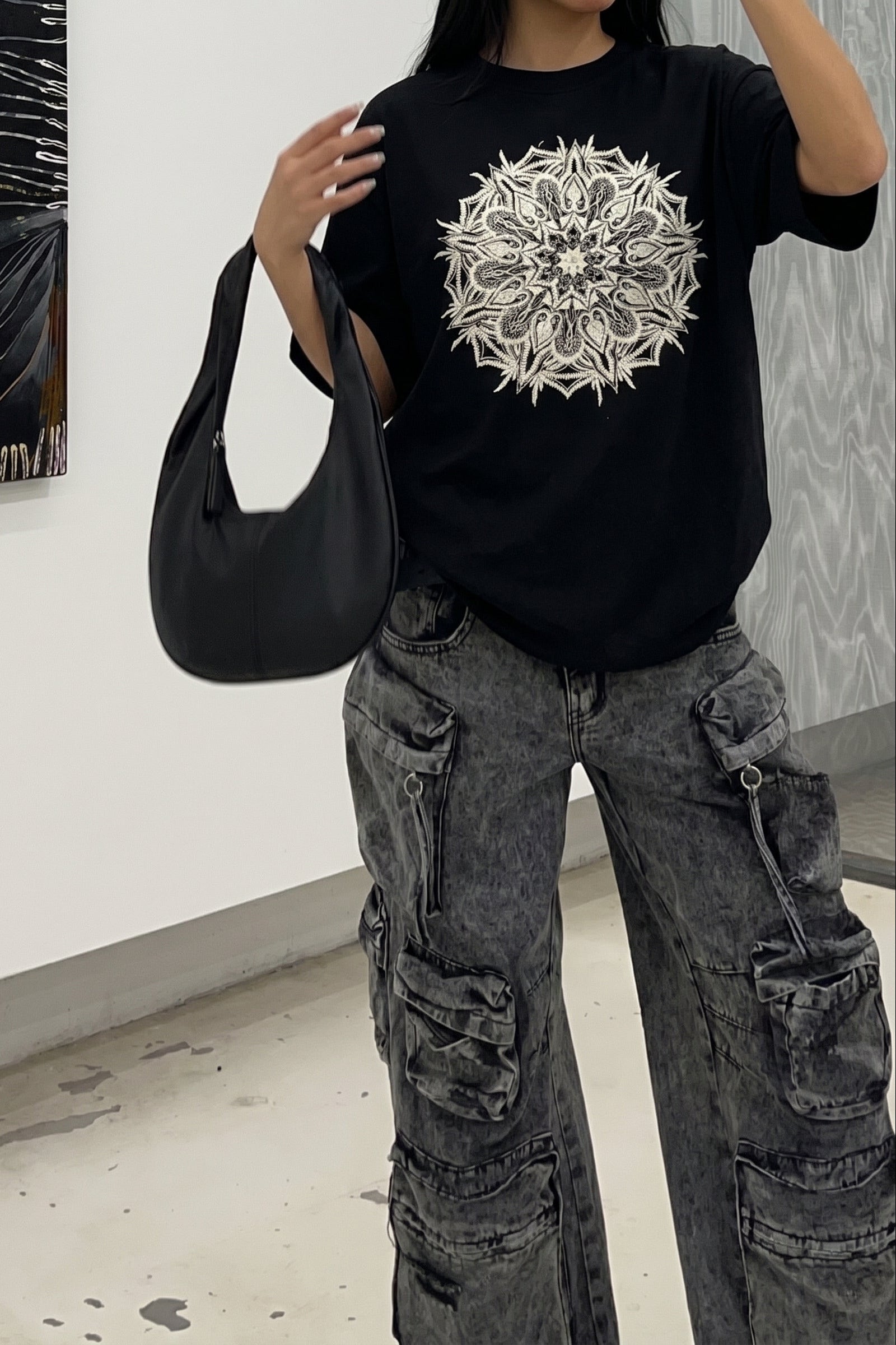 Person wearing a black t-shirt with a white mandala design, holding a black bag in an indoor setting.